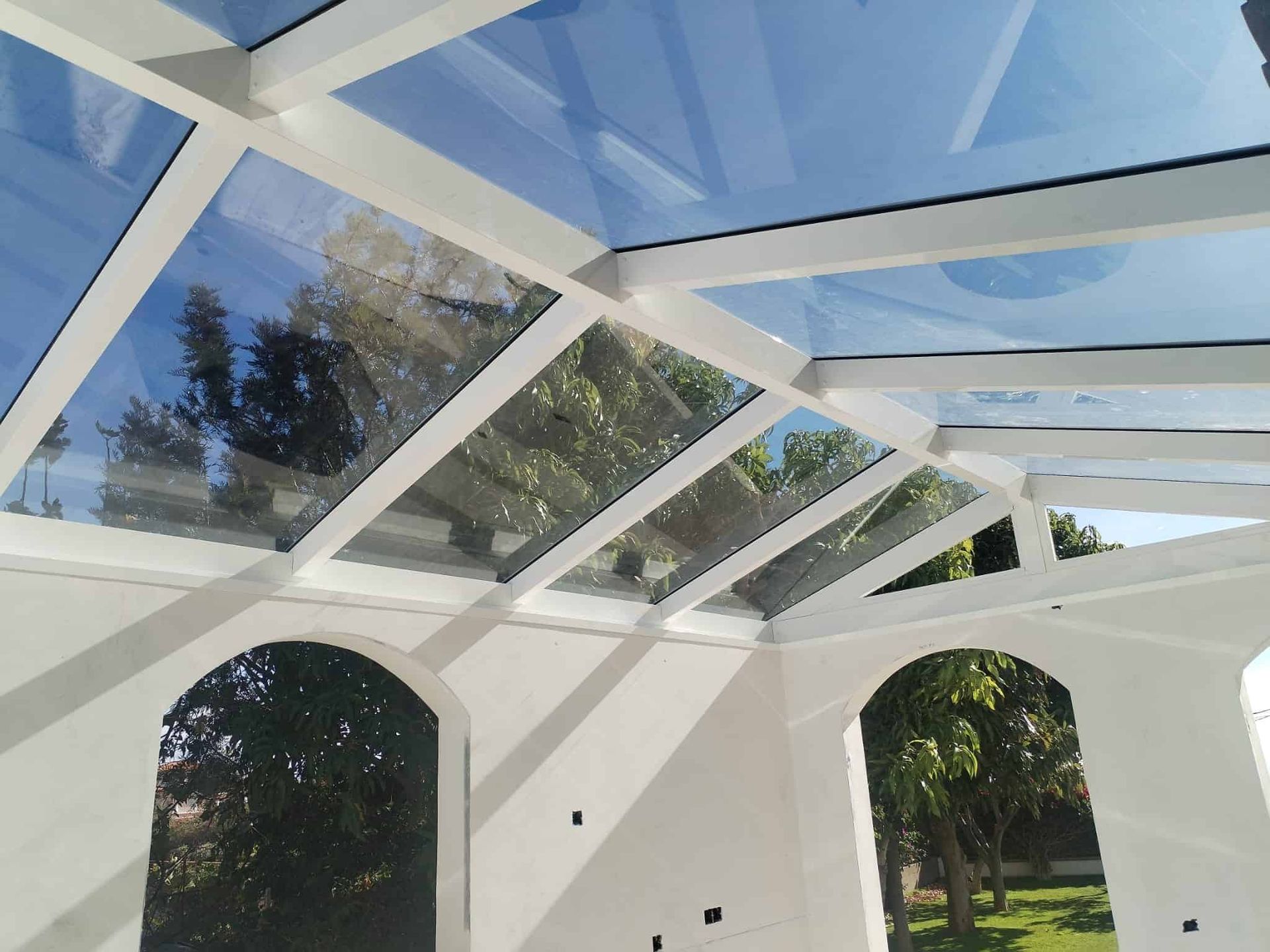 Techo de cristal con marco blanco, inclinado con paneles transparentes, que reflejan el cielo y los árboles. Paredes interiores blancas con aberturas arqueadas.