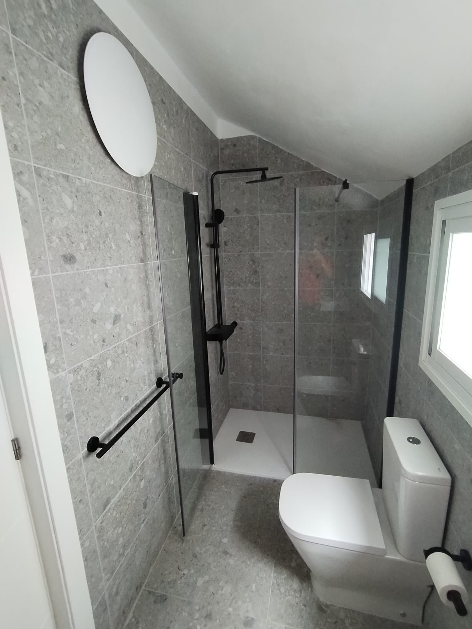 Baño con azulejos texturizados grises, ducha de cristal, accesorios negros, inodoro blanco y espejo redondo.