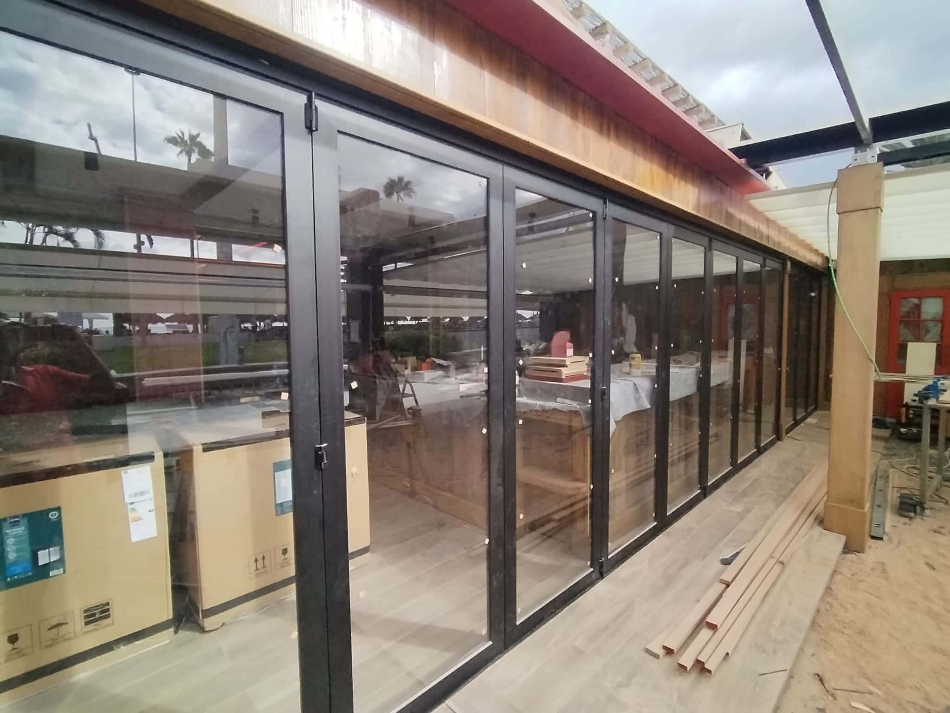 Puertas plegables de vidrio con marco negro, parcialmente abiertas, sobre un patio de madera, con techo de madera y cajas en el interior.