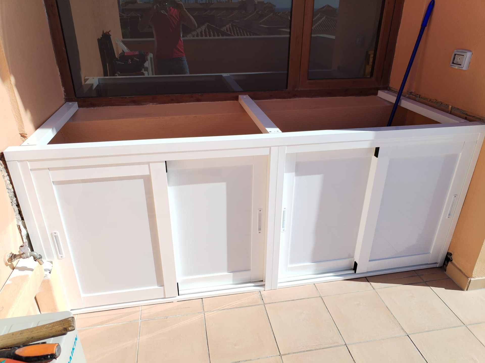 Armarios de almacenaje blancos empotrados en la pared de un balcón con puertas corredizas, frente a una ventana.