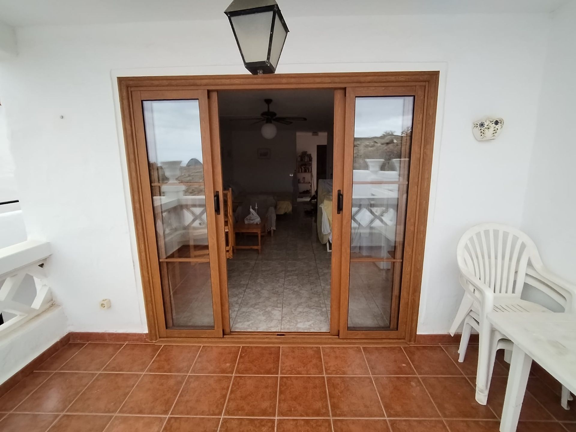 Unas puertas correderas con marco marrón dan paso a una sala de estar. Un balcón con baldosas de terracota cuenta con una silla y una mesa blancas.