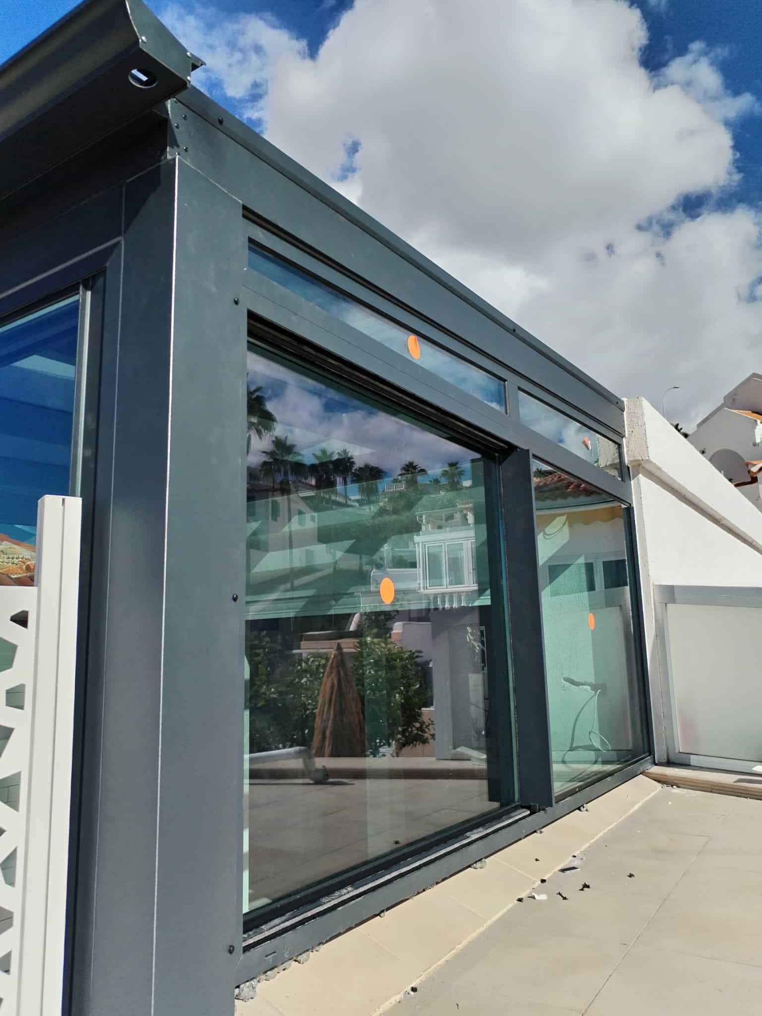 Ventanas y techo de vidrio con marco gris que reflejan un paisaje contra un cielo azul.