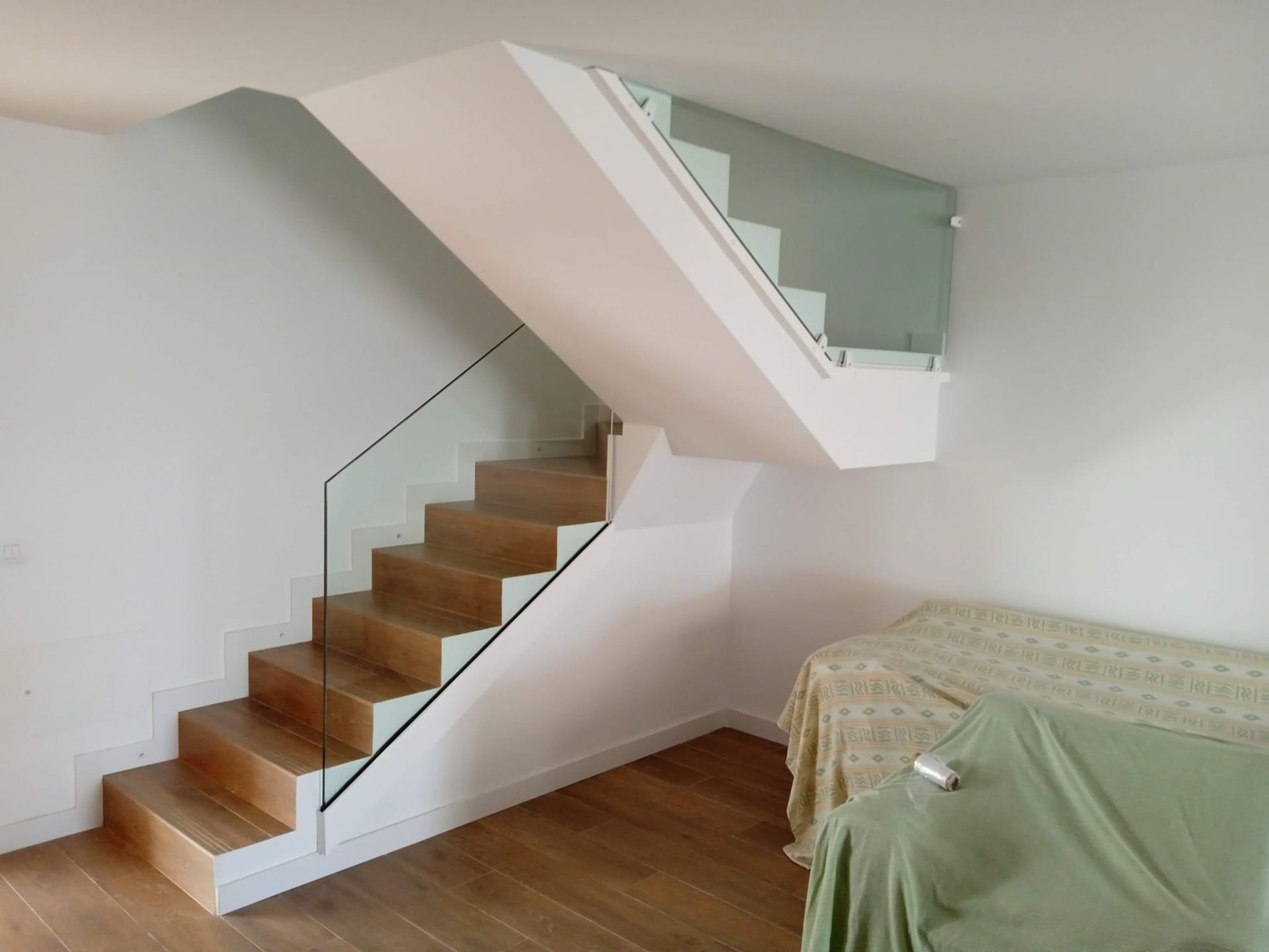 Escaleras de madera con barandilla de cristal en una habitación de paredes blancas, junto a muebles revestidos.