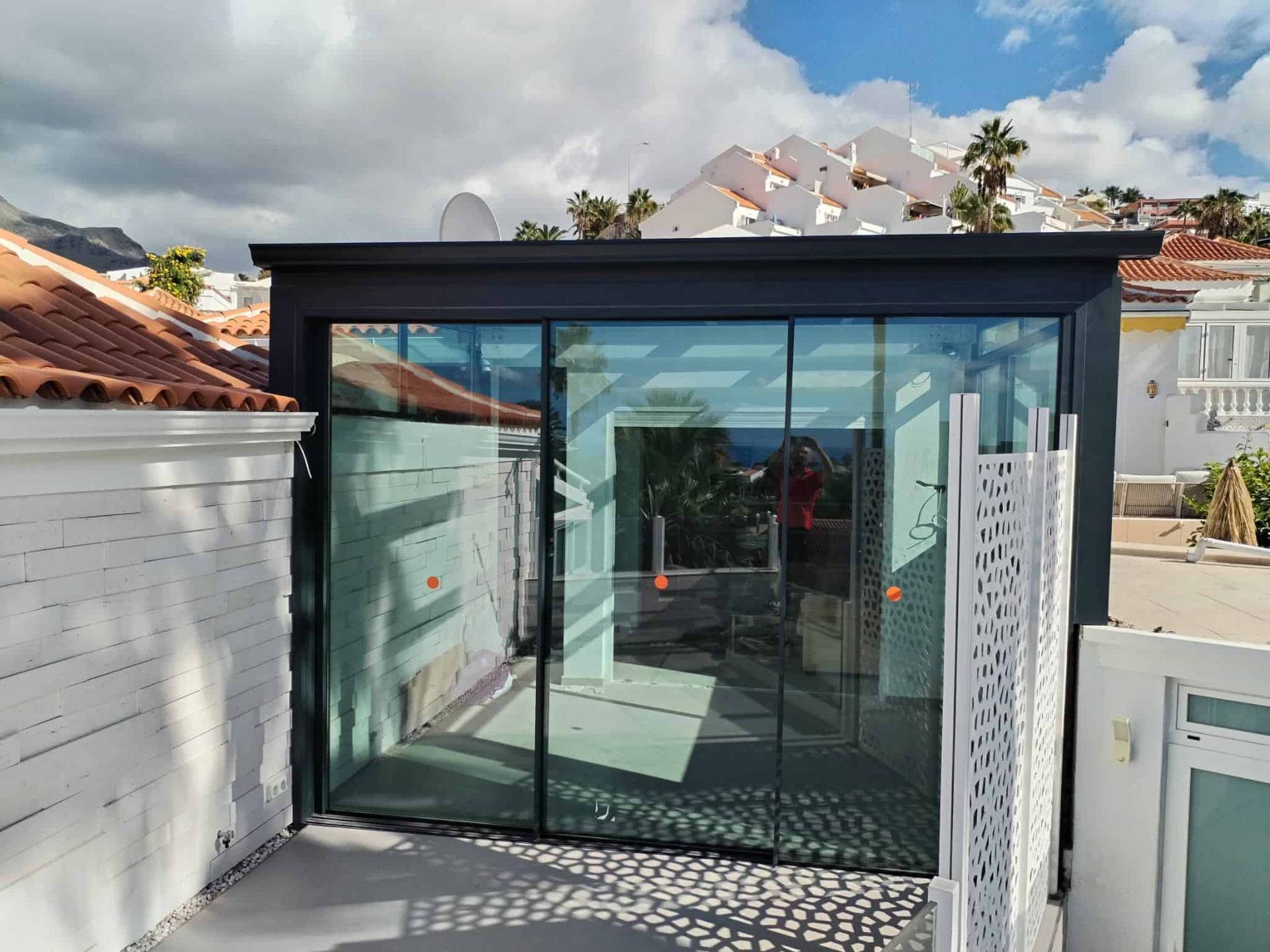 Terraza acristalada con marco negro, junto a una pared blanca y una valla estampada. Con vistas a un cielo parcialmente nublado.