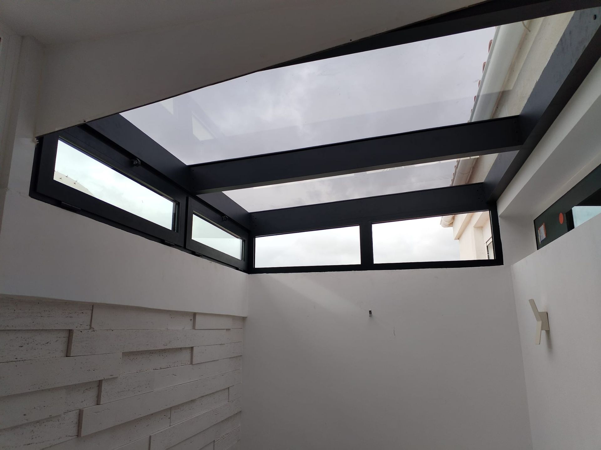 Tragaluz con marco de metal oscuro y paneles transparentes colocados en un hueco de pared blanca.