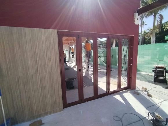 Se están instalando puertas plegables de vidrio con marco rojo; una persona las está arreglando. A la izquierda hay una pared texturizada.