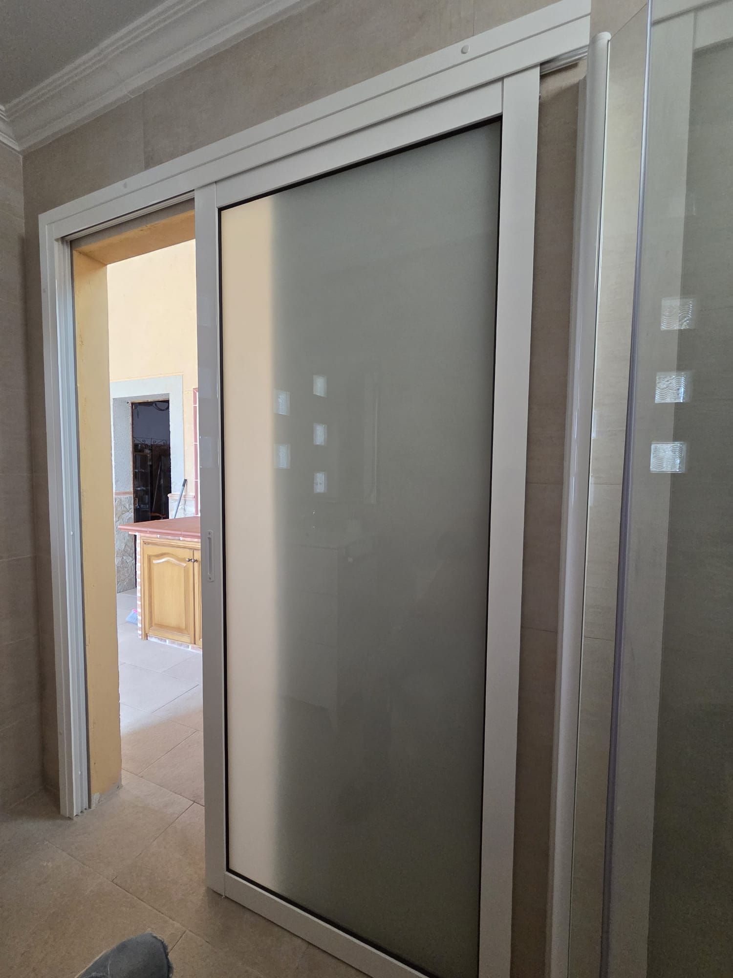 Puerta corrediza de vidrio esmerilado, marco blanco, pared de azulejos beige, puerta de entrada a otra habitación.