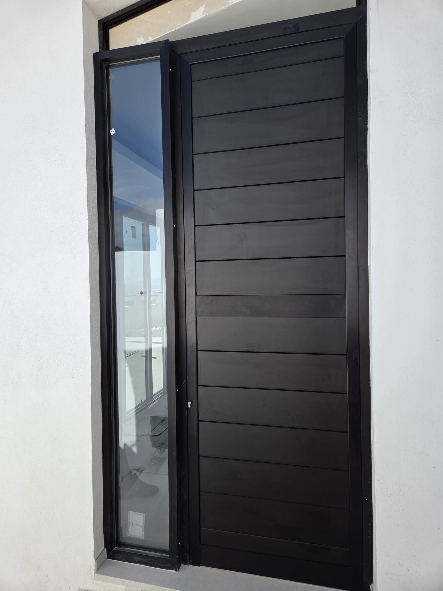 Puerta de entrada negra con un panel de vidrio lateral, colocada en una pared exterior blanca.