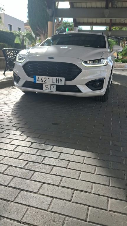 Taxis blancos circulando por una calle de la ciudad, con un carruaje tirado por caballos al fondo.