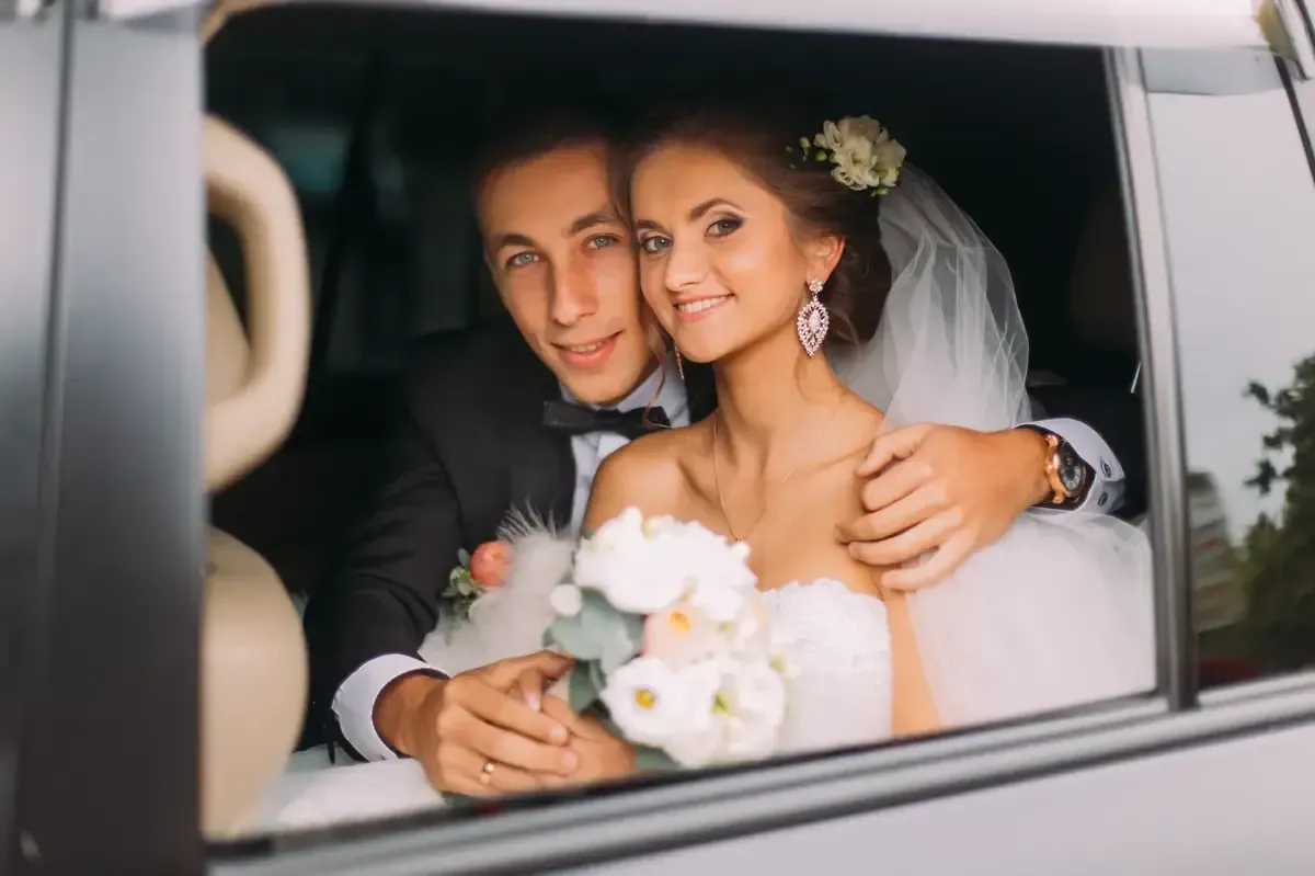 Recién casados ​​en un coche, sonriendo. La novia, vestida de blanco, sostiene el ramo. El novio, de esmoquin, la abraza.