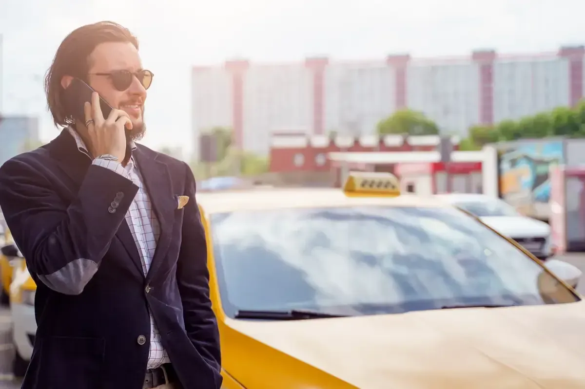 Hombre con traje y gafas de sol hablando por teléfono junto a un taxi amarillo en un entorno urbano.