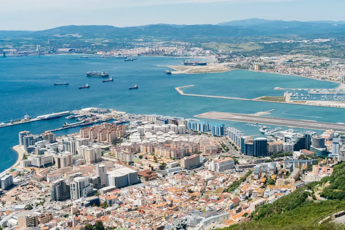 Vista aérea de Gibraltar, una ciudad costera, con edificios y puerto en foco, el mar y la costa española en el fondo.
