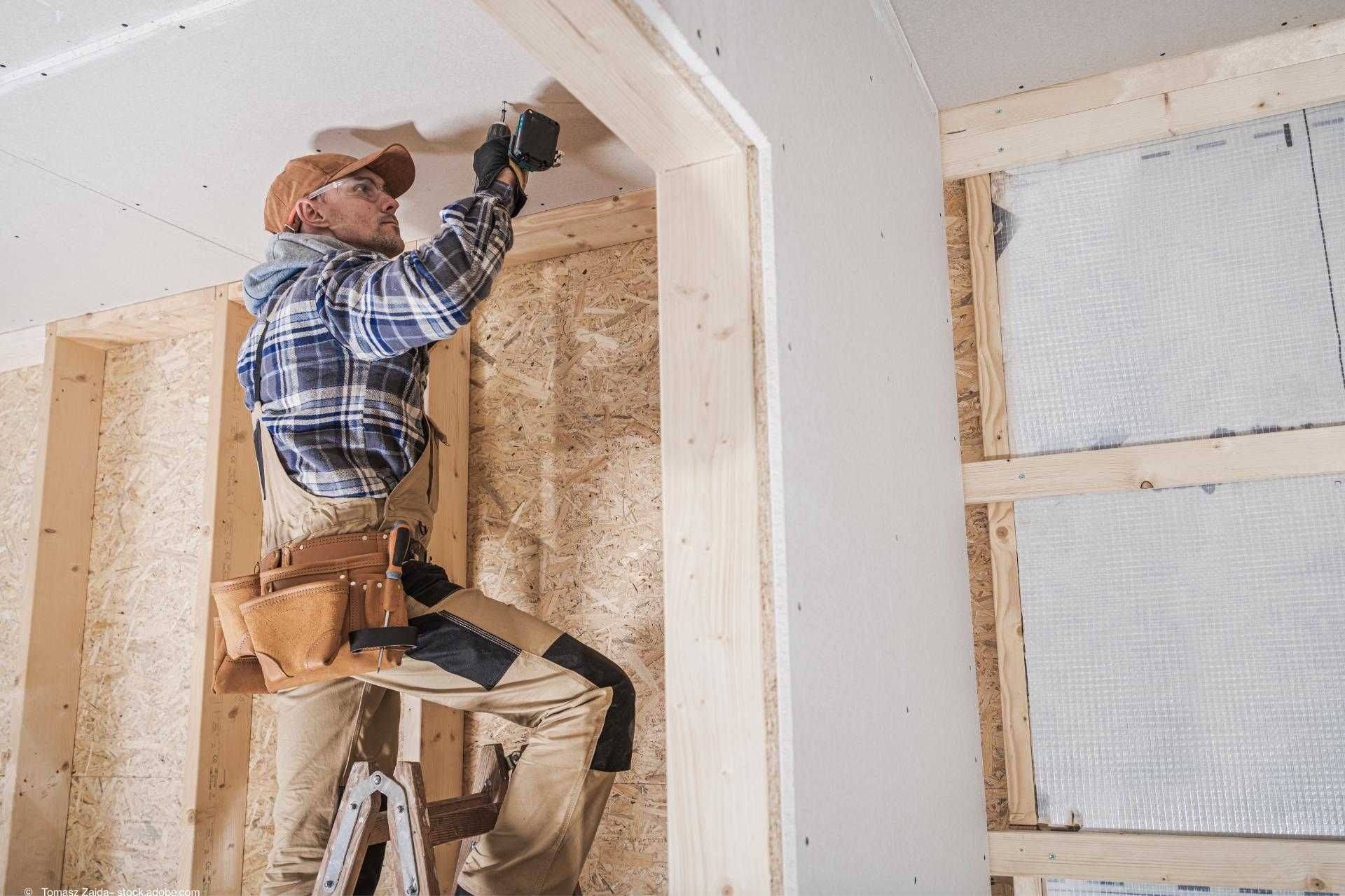 Handwerker montiert Gipskartonplatte an Decke in einem Rohbau mit Holzständerwerk und OSB-Platten.