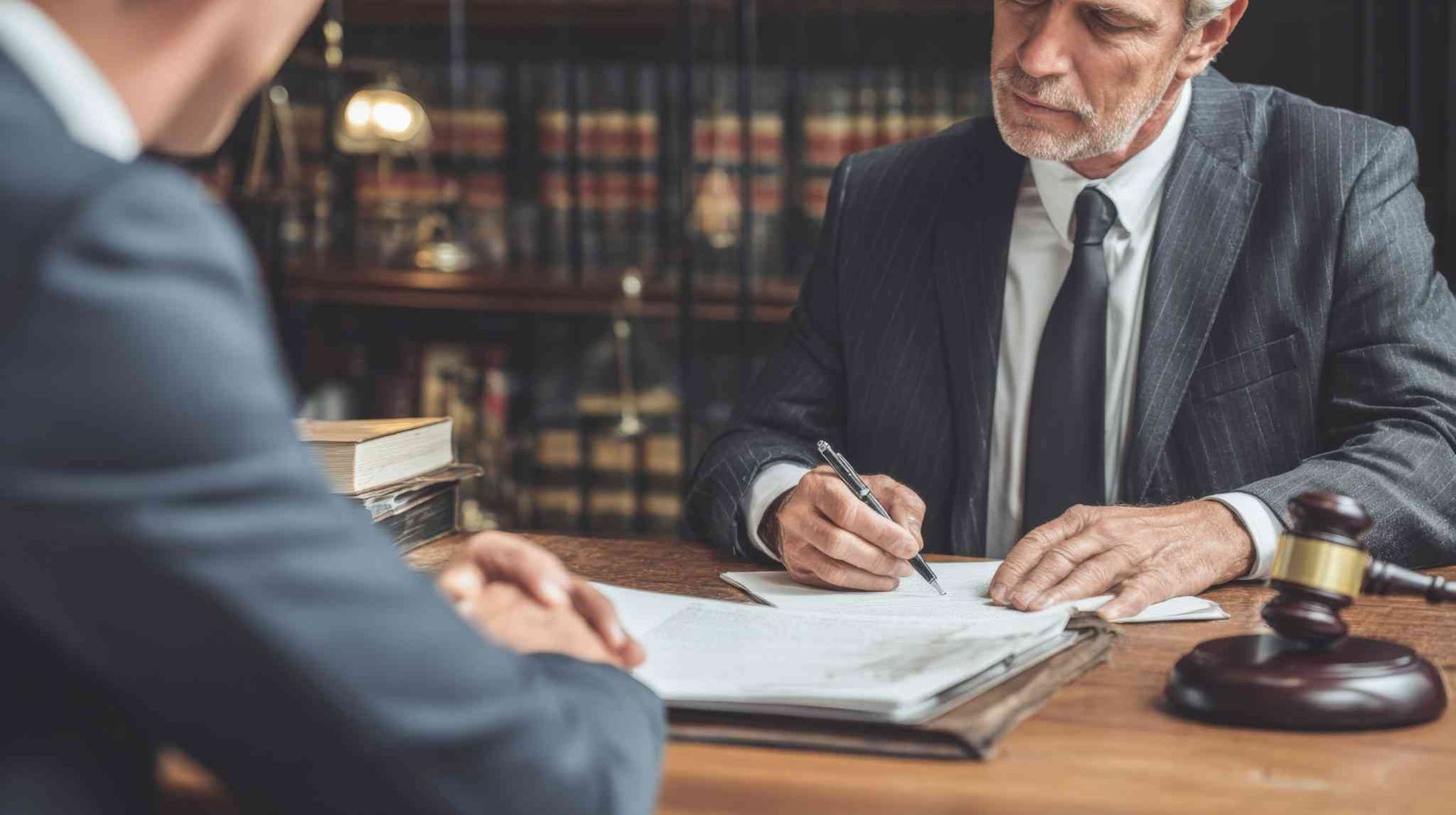 Un abogado de traje firma documentos en un escritorio de madera con un mazo, mientras un cliente está sentado.