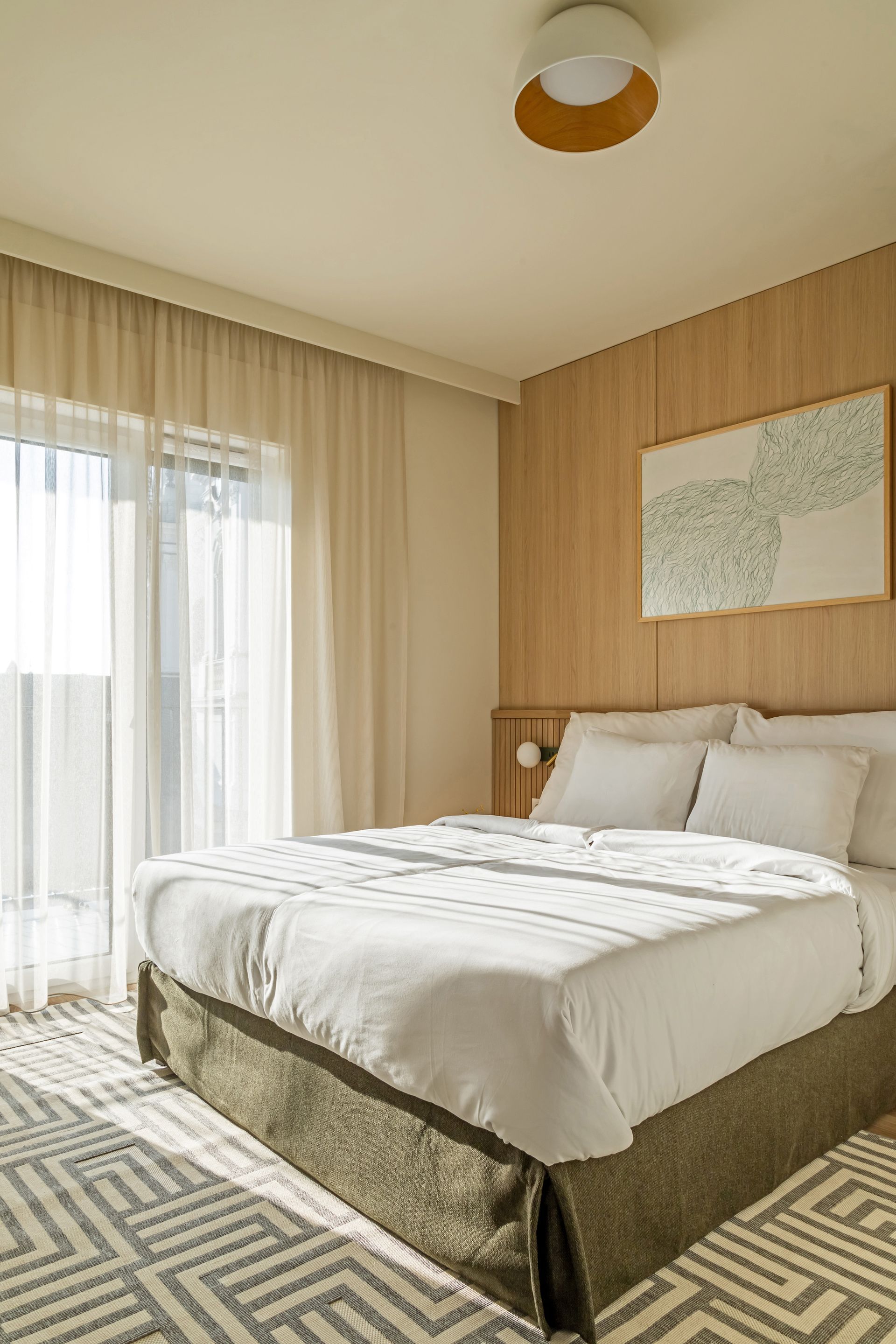 Bright bedroom with a large bed, sheer curtains, wooden headboard, and patterned rug in Marone Suites Budapest.