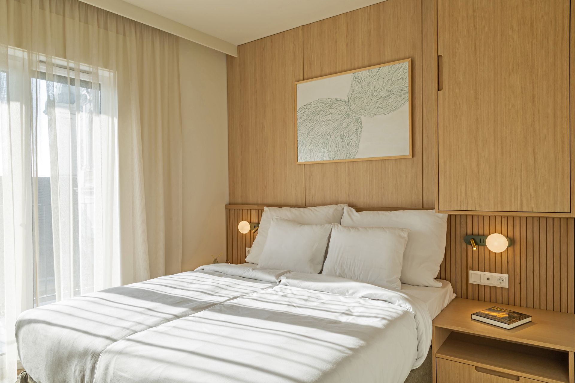 Bedroom with light wood paneling, a bed with white linens, artwork, and a balcony door with sheer curtains in Marone Suites.