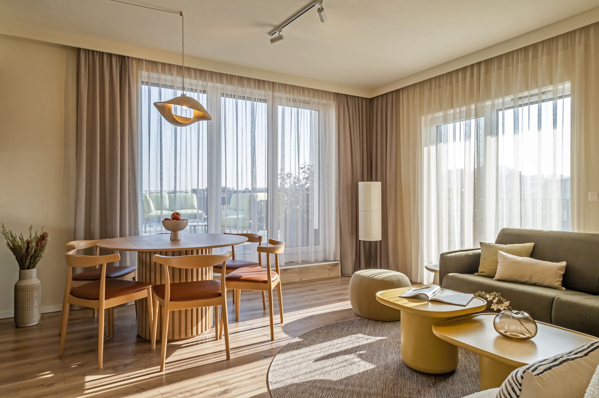 Sunny living room with dining table, sofa, sheer curtains, glass doors leading to a rooftop in neutral tones. Marone Suites