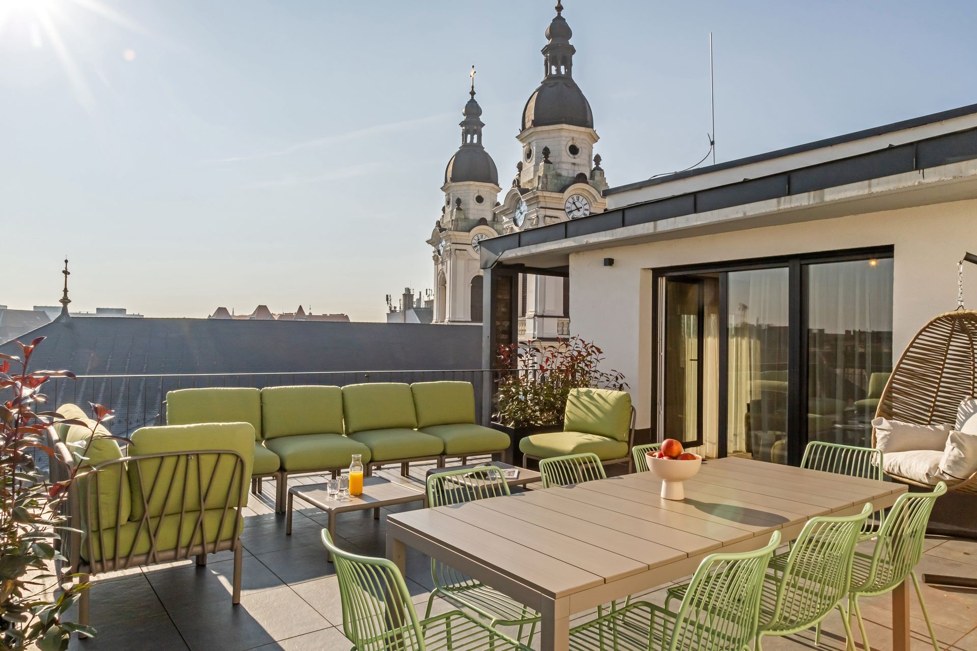 Rooftop patio with green furniture, dining table, and city views, with church spires in the background in Marone Suites.