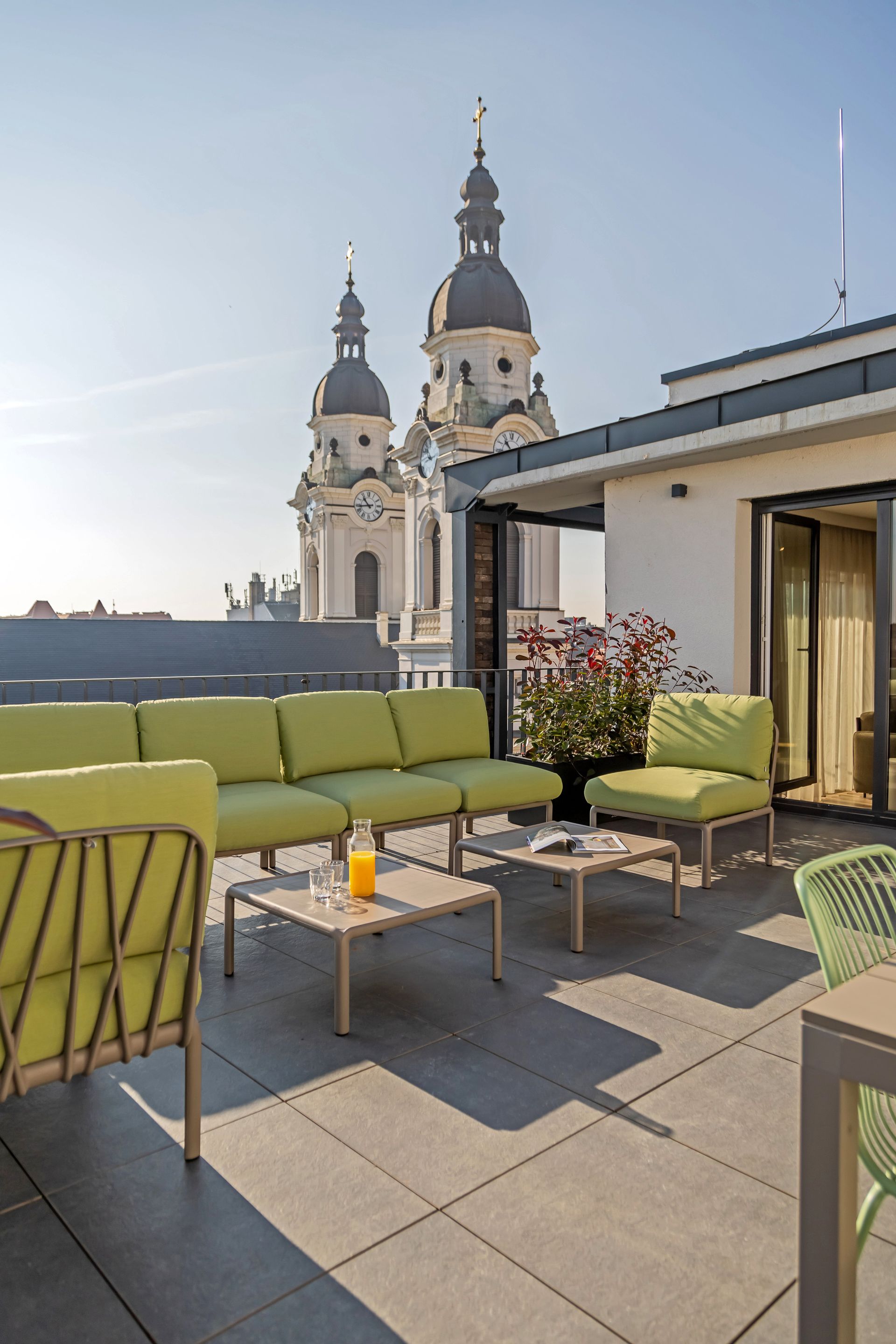 Rooftop patio with green furniture, small tables, and views of a church with two domes in Marone Suites Budapest.