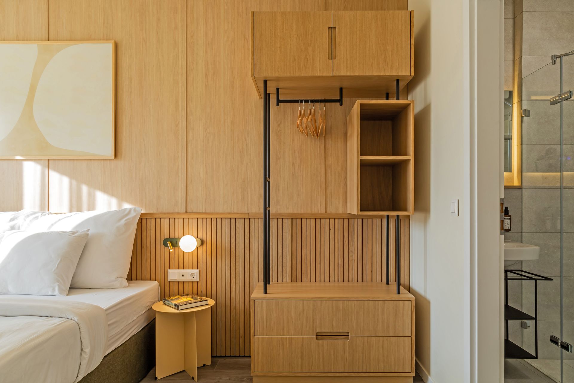 Bedroom with light wood paneling, bed, wardrobe, a small side table and a door opening to an en-suite bathroom. Marone Suites