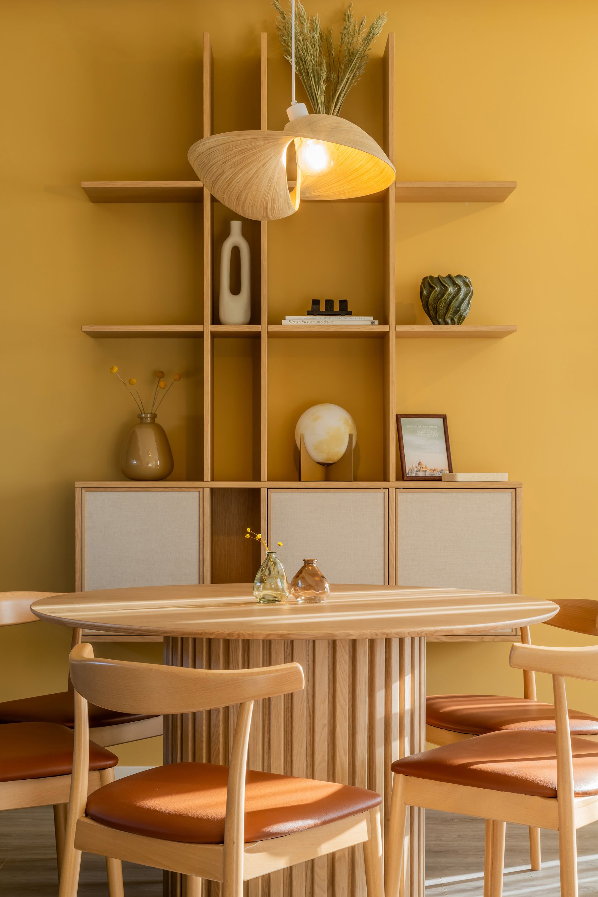 Dining area with yellow wall, wooden table, chairs, and decorative shelf with items in Marone Suites Budapest.