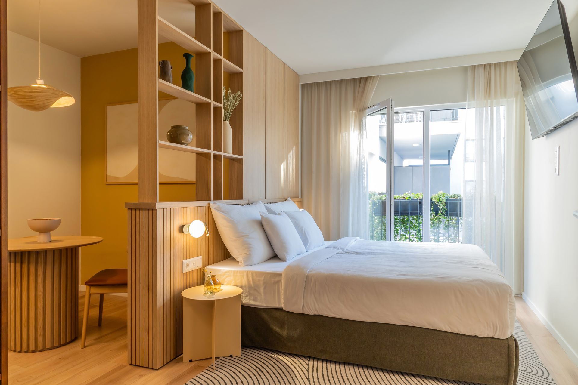 Studio apartment with wooden built-ins, bed with white bedding, small round table, and a window in Marone Suites Budapest.