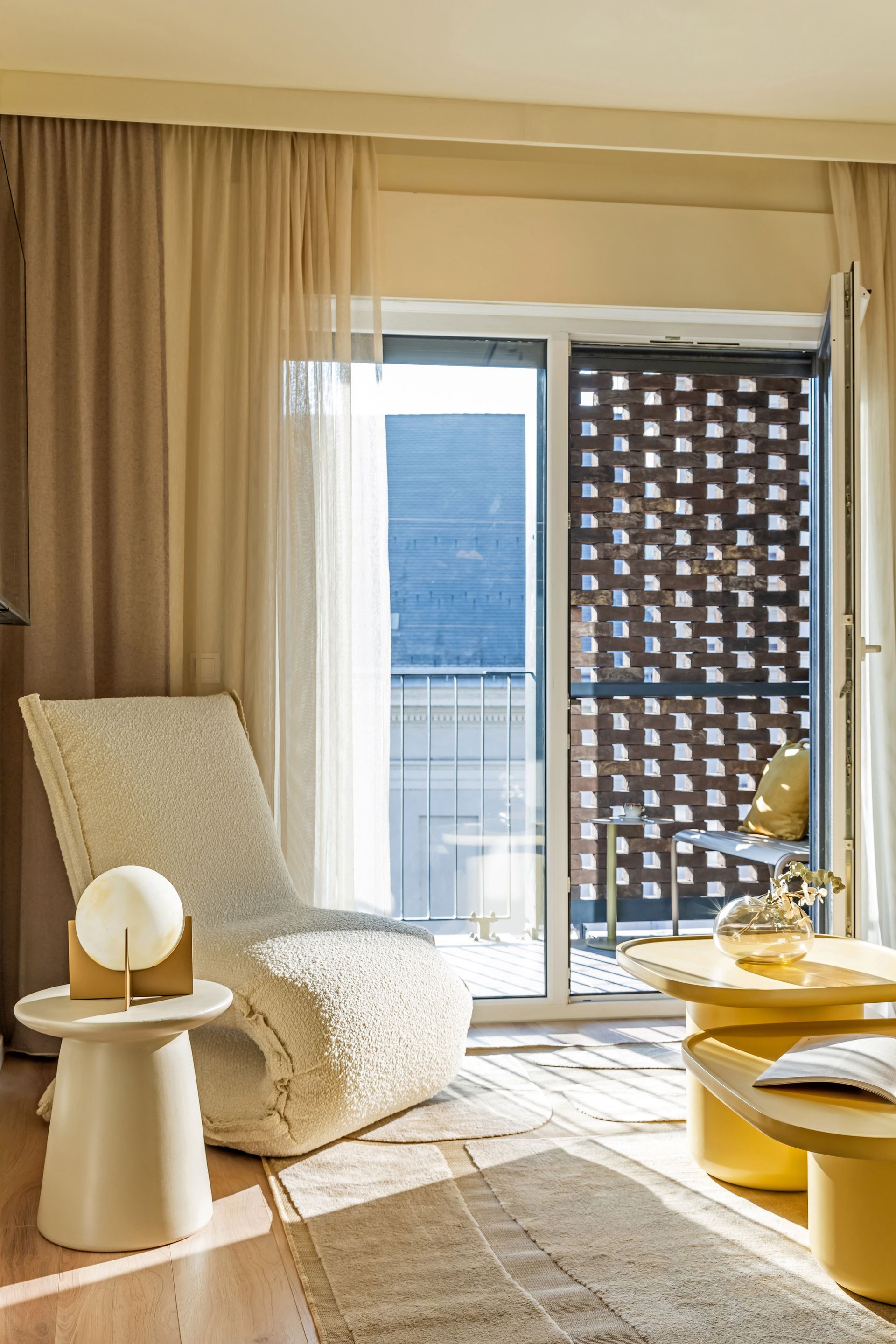 Cozy living room with cream lounge chair, small table with lamp, and balcony. Sunny light, neutral tones in Marone Suites.