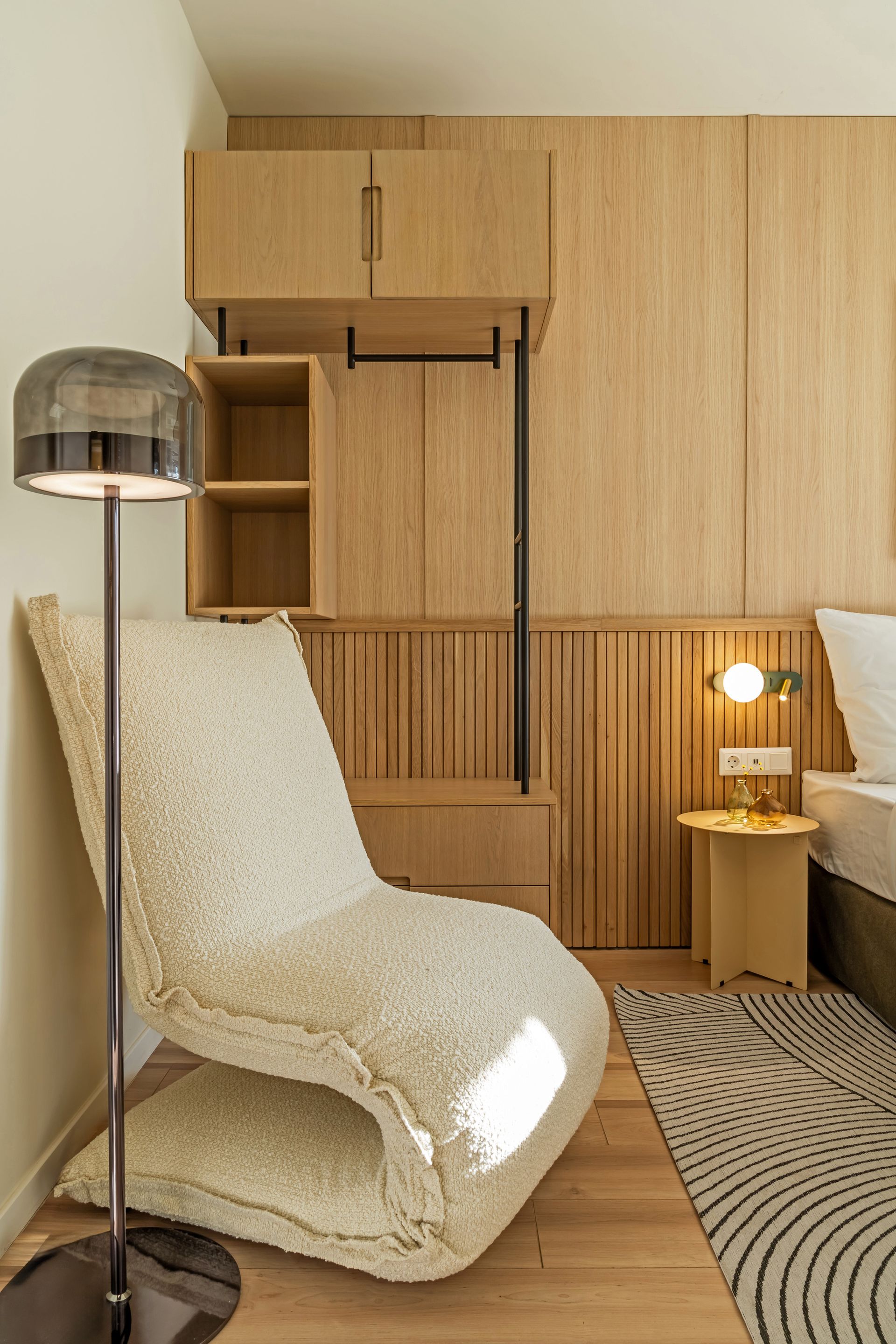Cozy bedroom corner with a fluffy white chair, wooden paneling, and a floor lamp in Marone Suites Budapest.