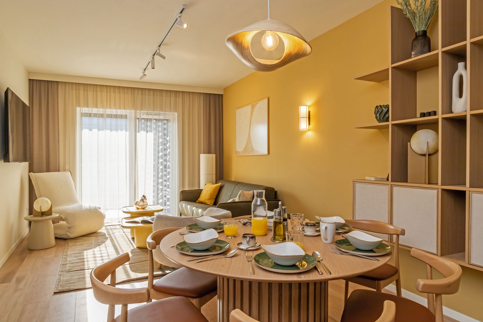 Cozy dining and living room with yellow and beige wall, round table set for four, and open shelving in Marone Suites.
