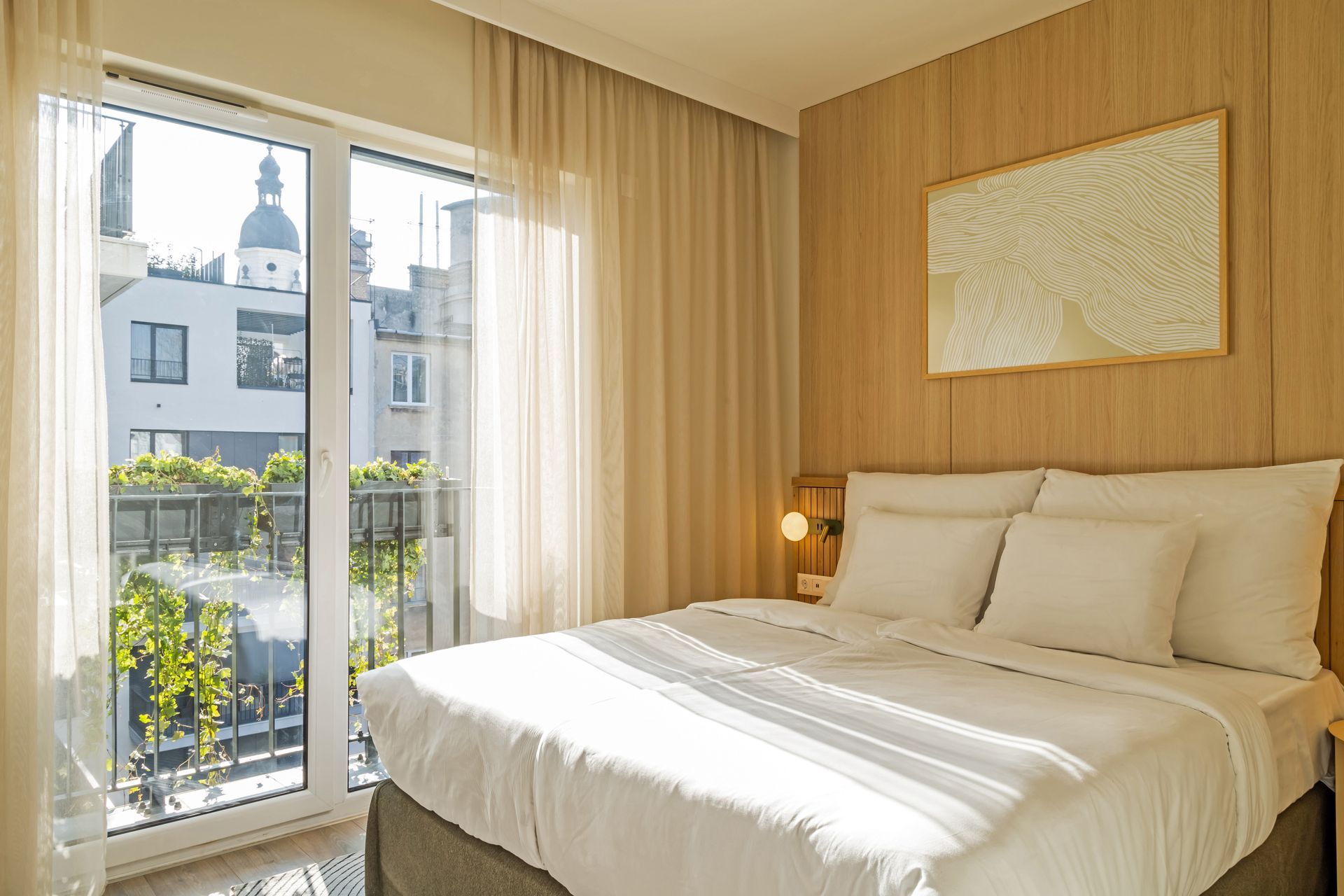Bedroom with a double bed, large window, sheer curtains, light wood paneling, artwork and garden view in Marone Suites.