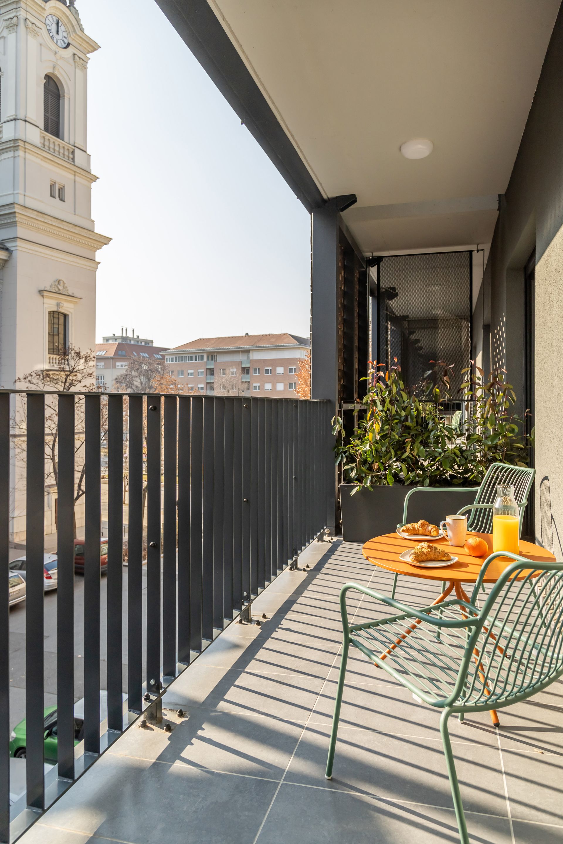 Balcony overlooking a city with table set for breakfast; chairs, sunlight, view of church.