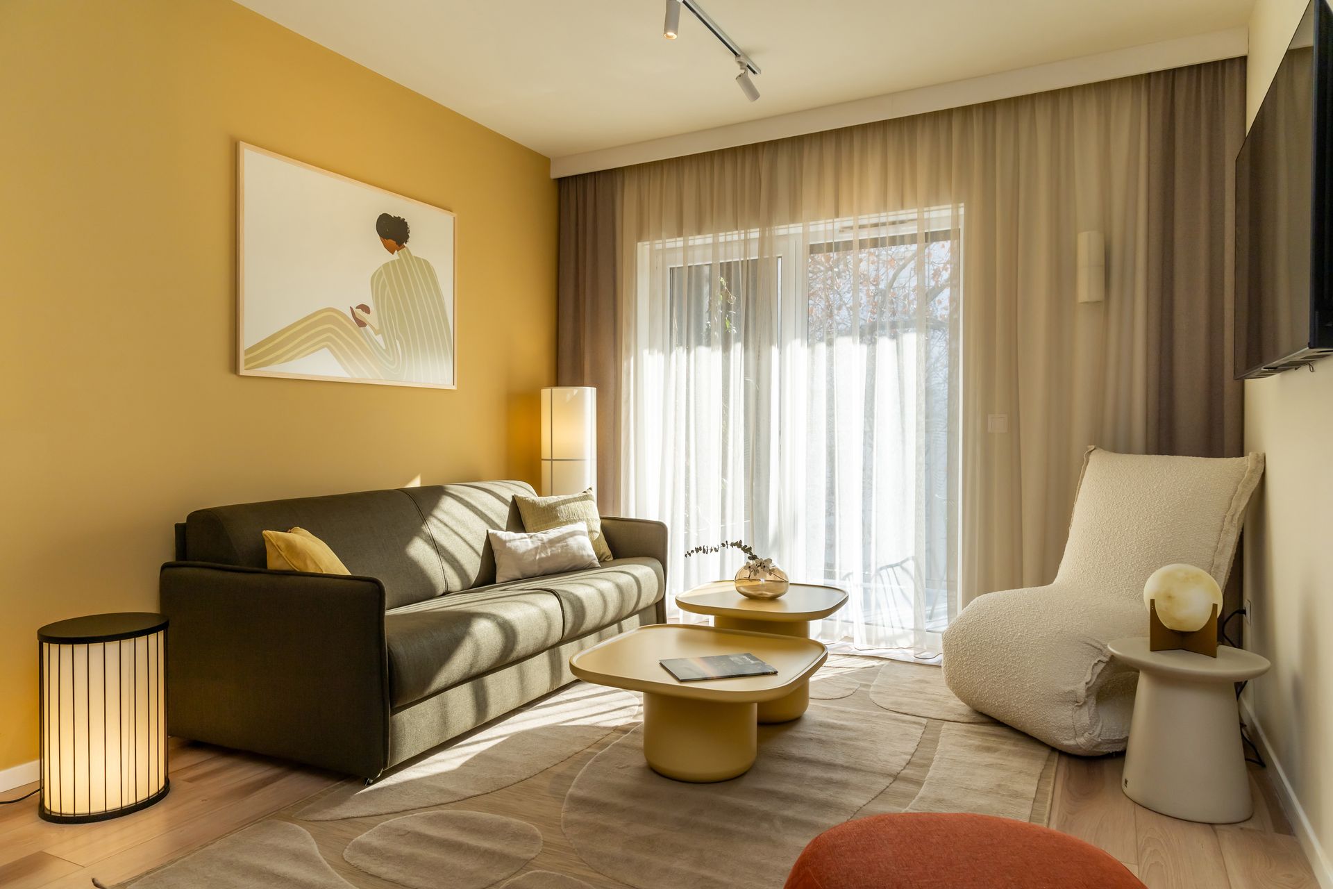 Sunny living room with olive sofa, yellow wall, beige rug, and large windows open to a balcony in Marone Suites Budapest.