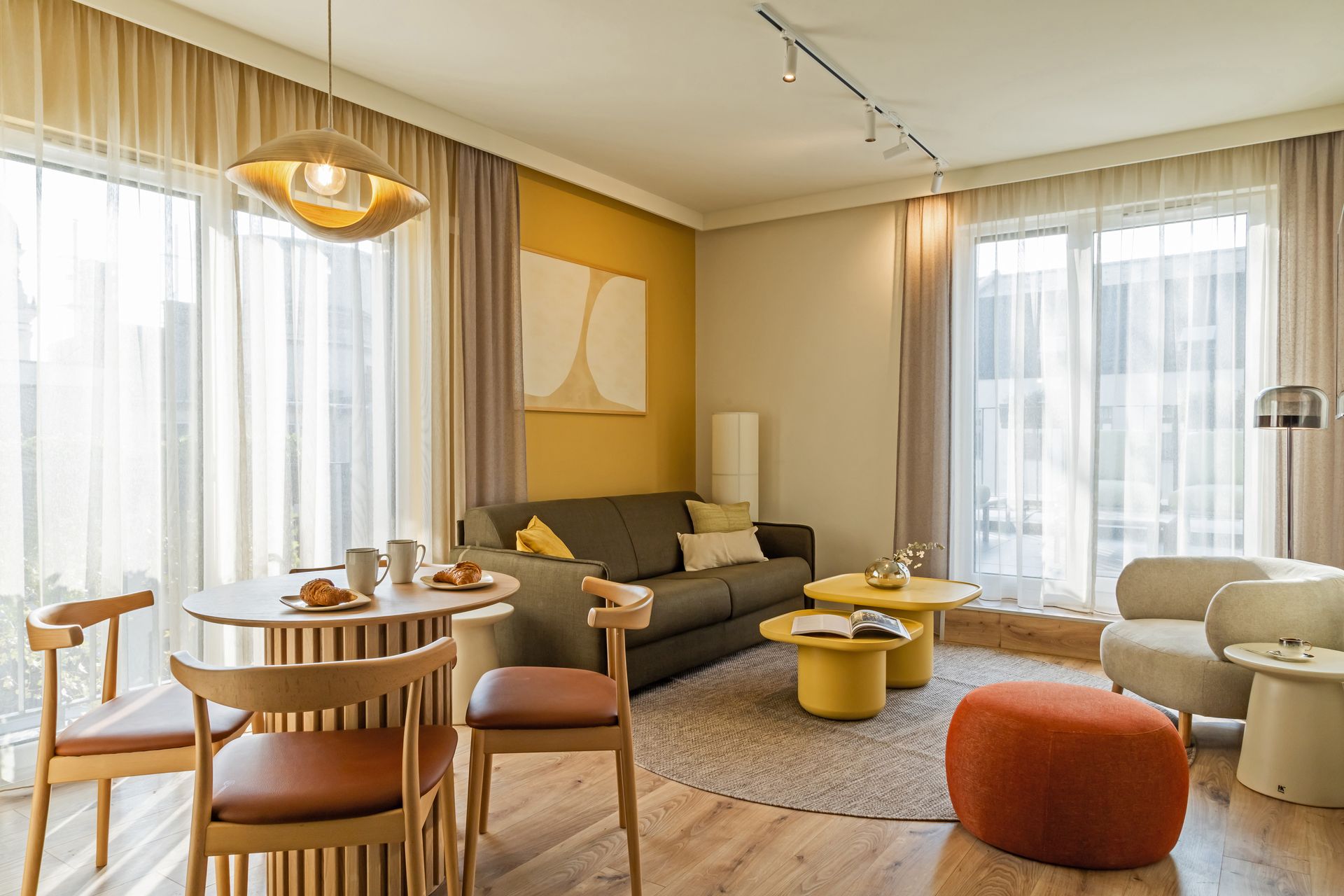 Sunny living room with sofa, round table, chairs, yellow coffee tables, and large doors leading to a balcony. Marone Suites