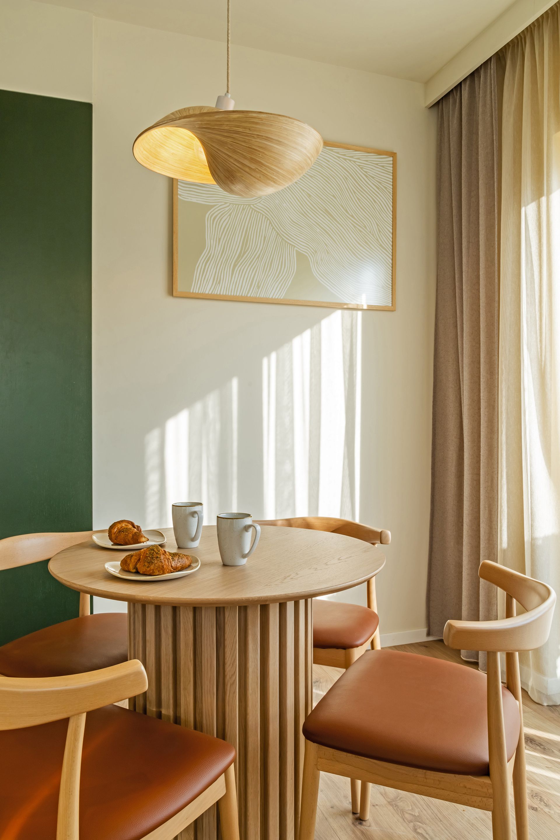 Dining area with round wooden table, chairs, art and hanging light fixture. Sunlight streams through a window. Marone Suites