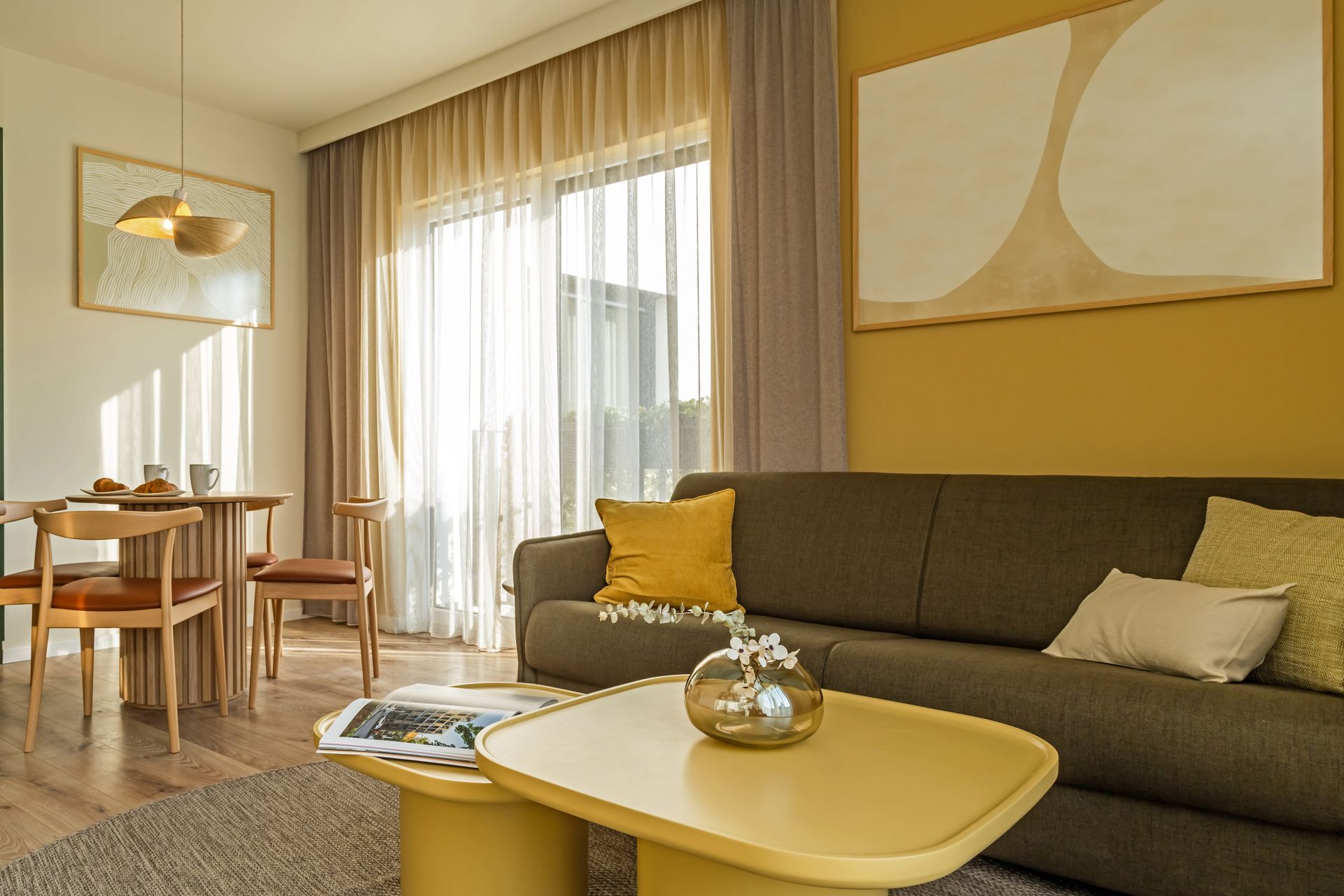 Cozy living room with yellow accent wall, olive green sofa, round table, lots of light and a balcony in Marone Suites.
