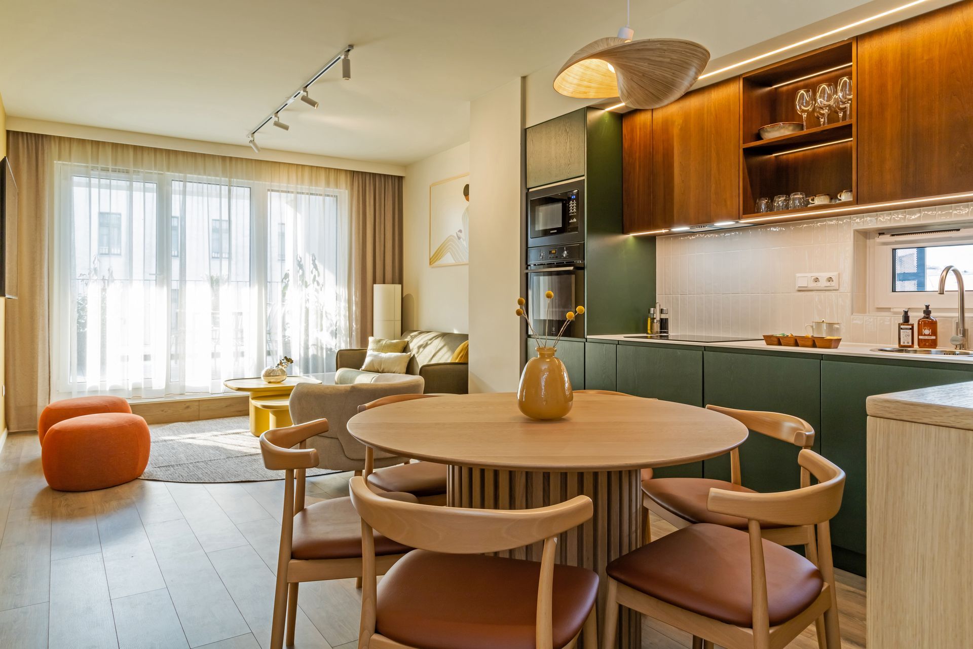 Open-concept kitchen and dining area with green cabinets, wooden table, and living room in the background in Marone Suites.