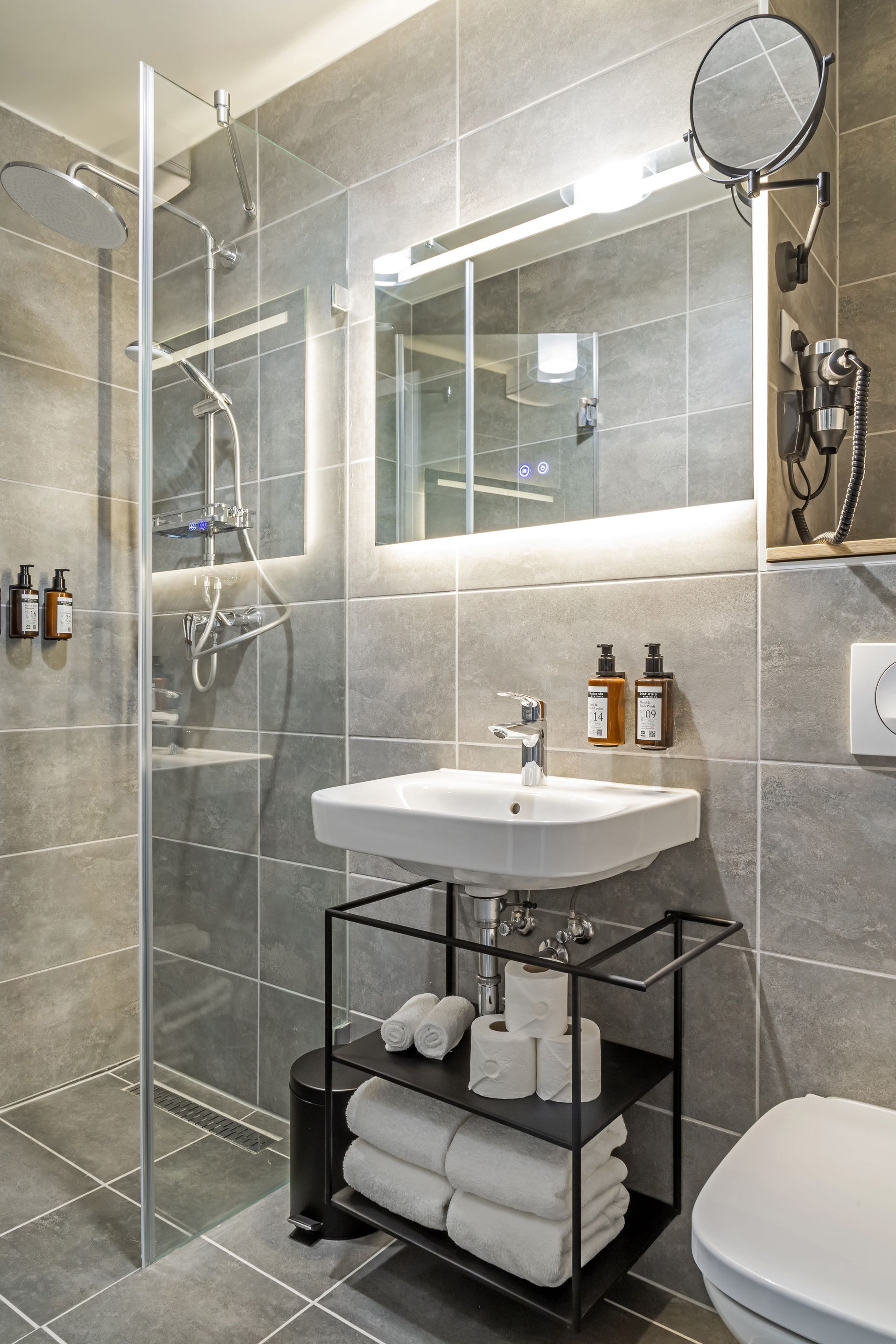 Modern bathroom with gray tile, walk-in shower, sink, mirror, toiletries and towels in Marone Suites Budapest.