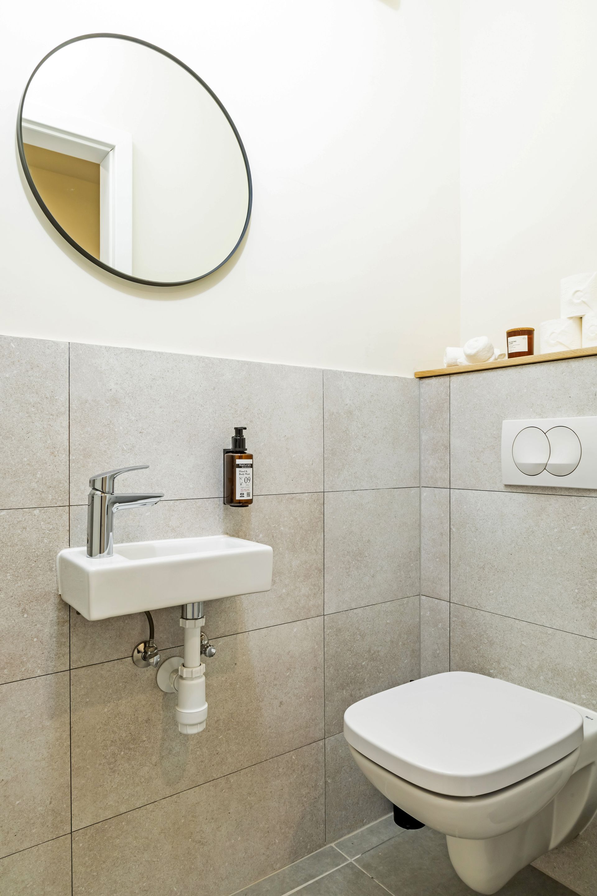 Small modern toilet with white walls, gray tile, round mirror, and wall-mounted toilet and sink in Marone Suites Budapest.