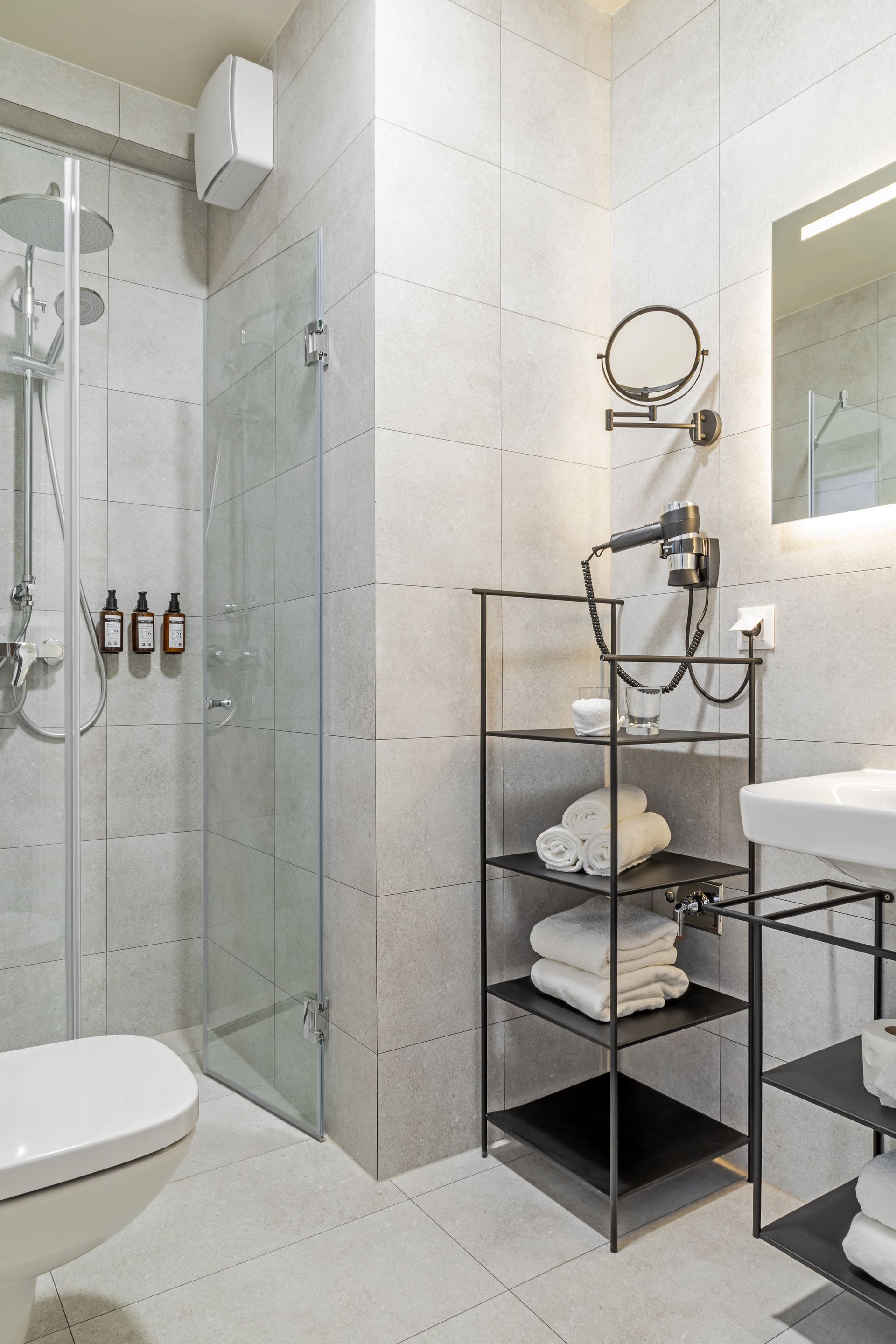 Modern bathroom with walk-in shower, toilet, shelving unit, sink, and mirror with illuminated lighting in Marone Suites.