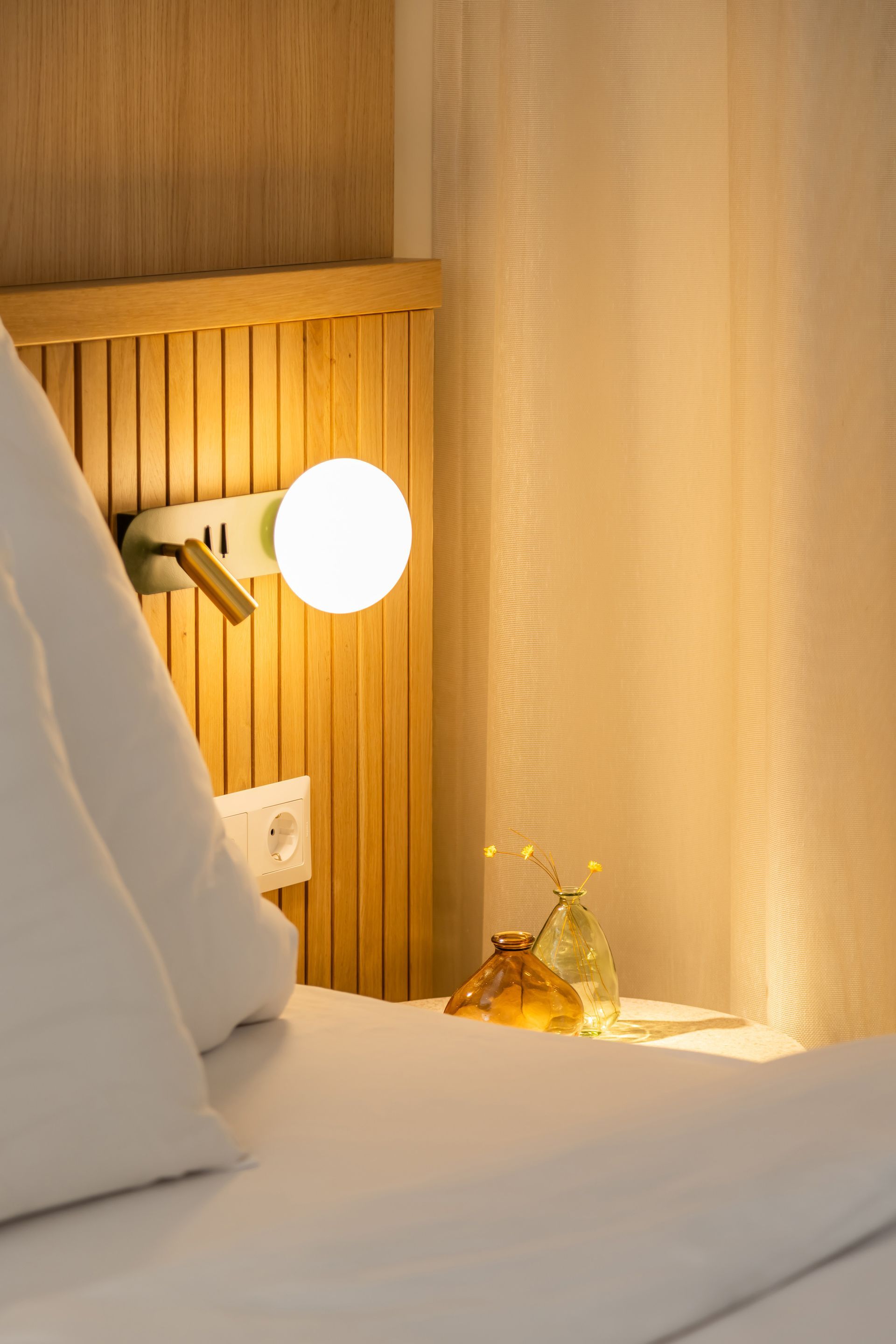 Bedside table with lamp and decorative vases, next to a bed with white pillows and a wooden headboard in Marone Suites.