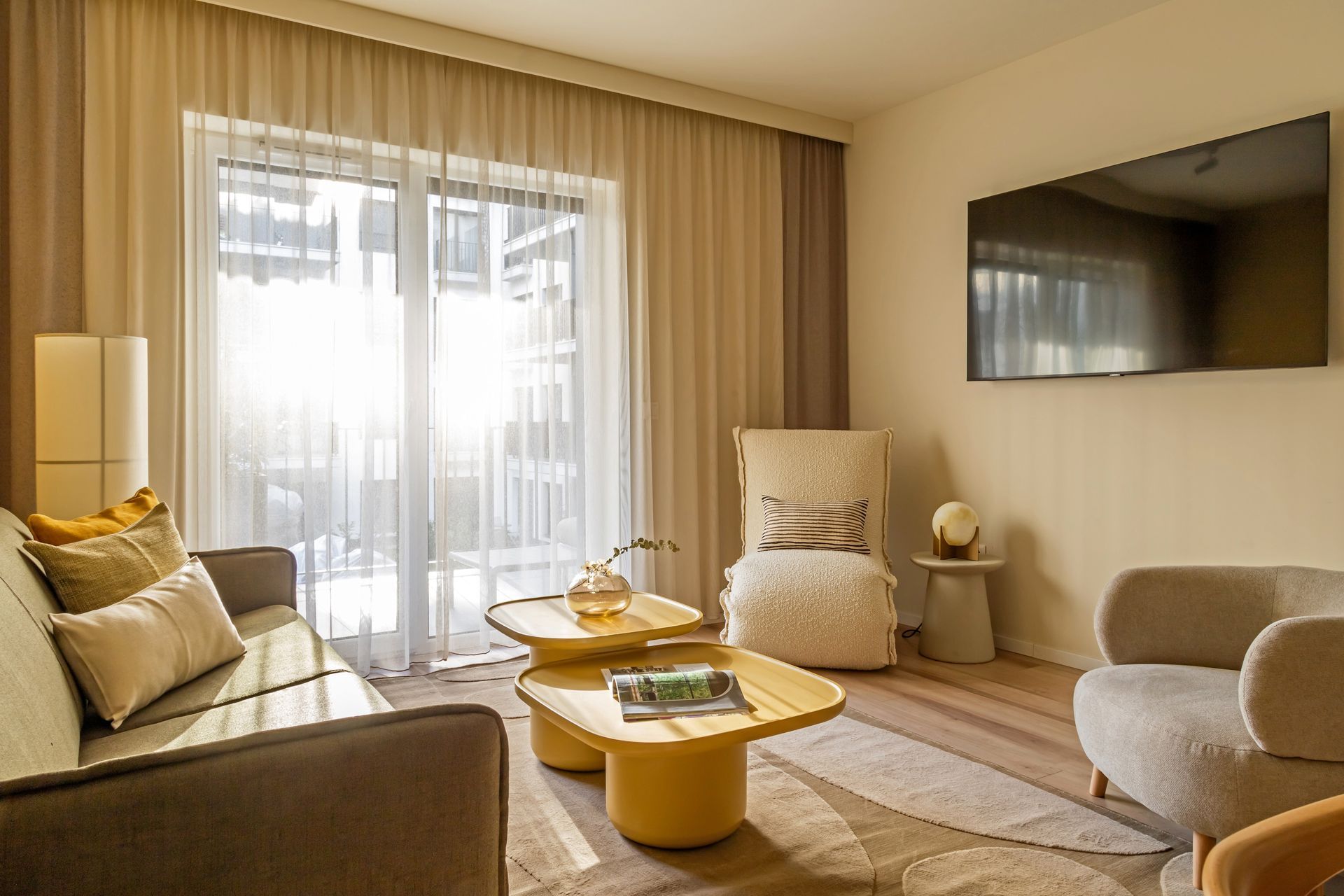 Living room with light beige walls, sofa, armchair, and a TV; a balcony is visible through the window in Marone Suites.