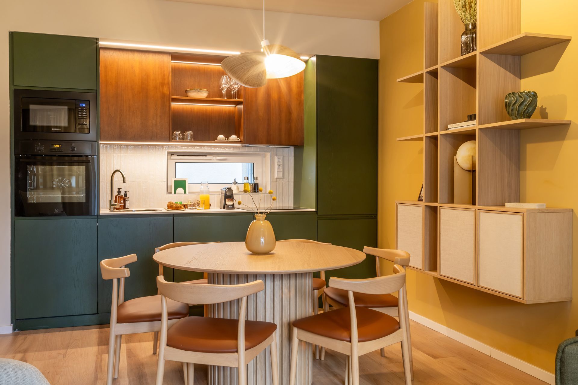 Kitchen with green and wood cabinetry, round table, chairs, and yellow wall with built-in shelving in Marone Suites Budapest.