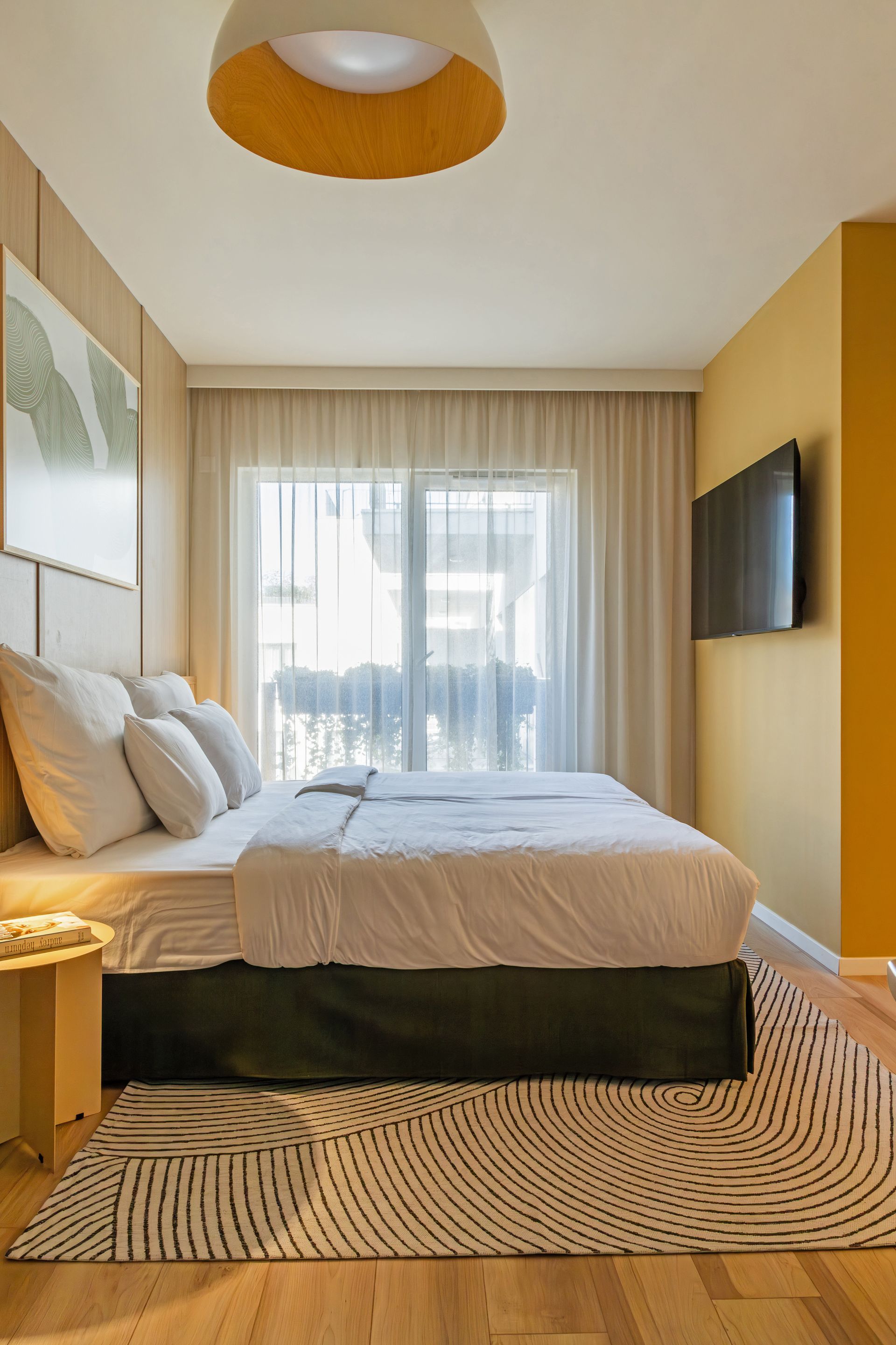 Studio apartment with a bed, TV, and a window. Yellow wall, white bedding, and patterned rug in Marone Suites Budapest.
