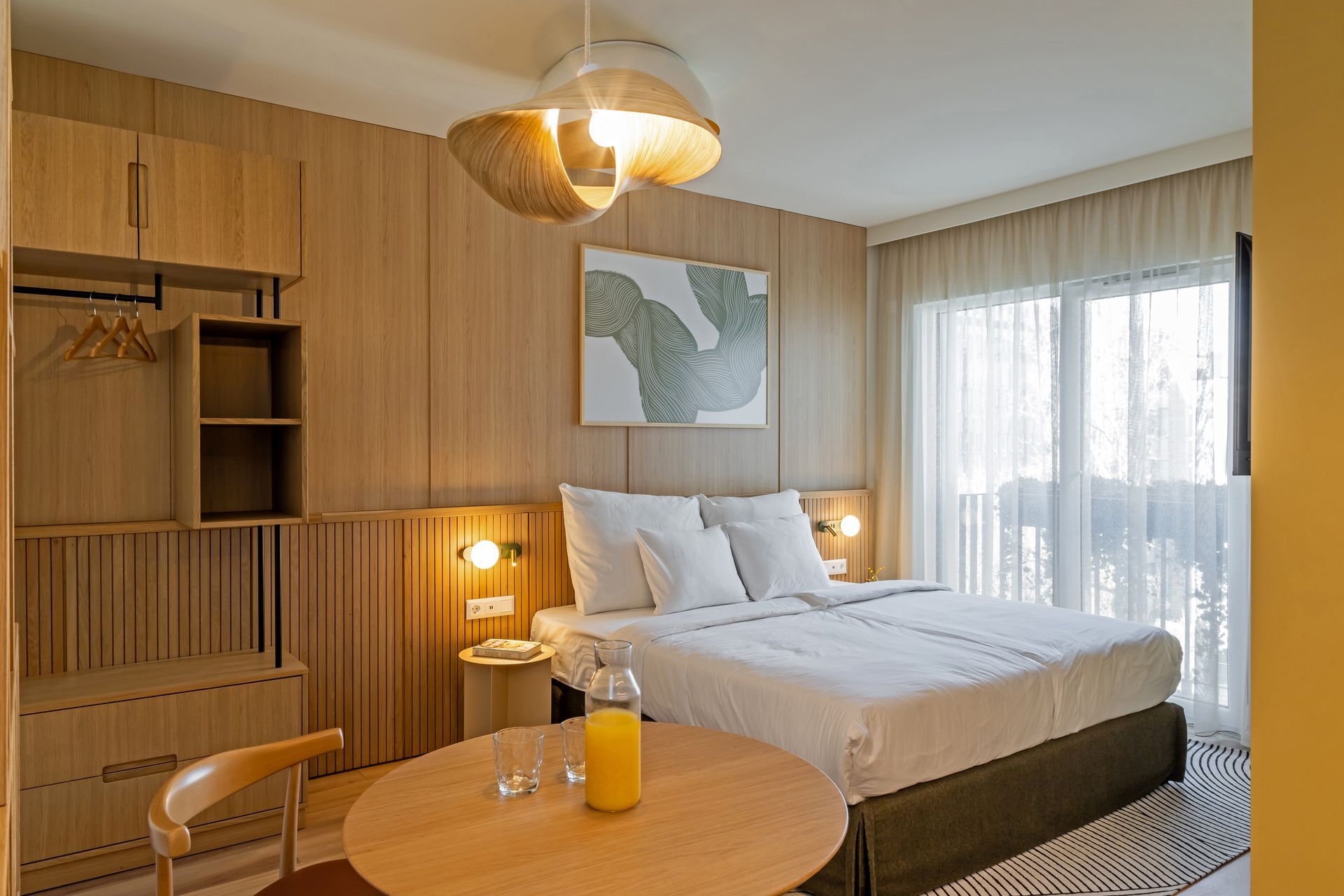 Studio apartment with a double bed, wooden furniture, artwork, and a table with drinks by a window in Marone Suites Budapest.