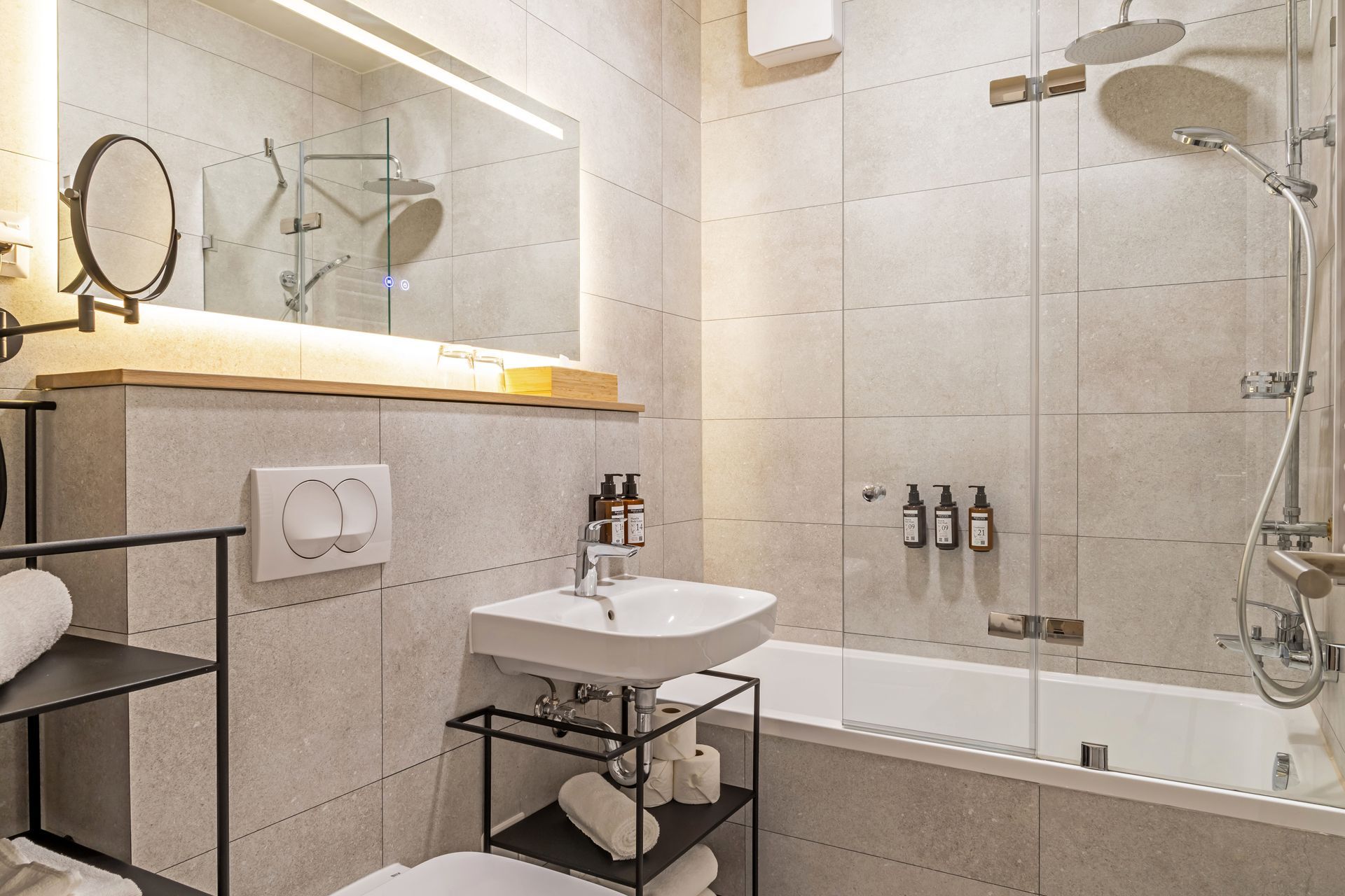 Modern bathroom with a bathtub, shower, sink, and mirror. Gray tiles, toiletries, white towels and shelving in Marone Suites.