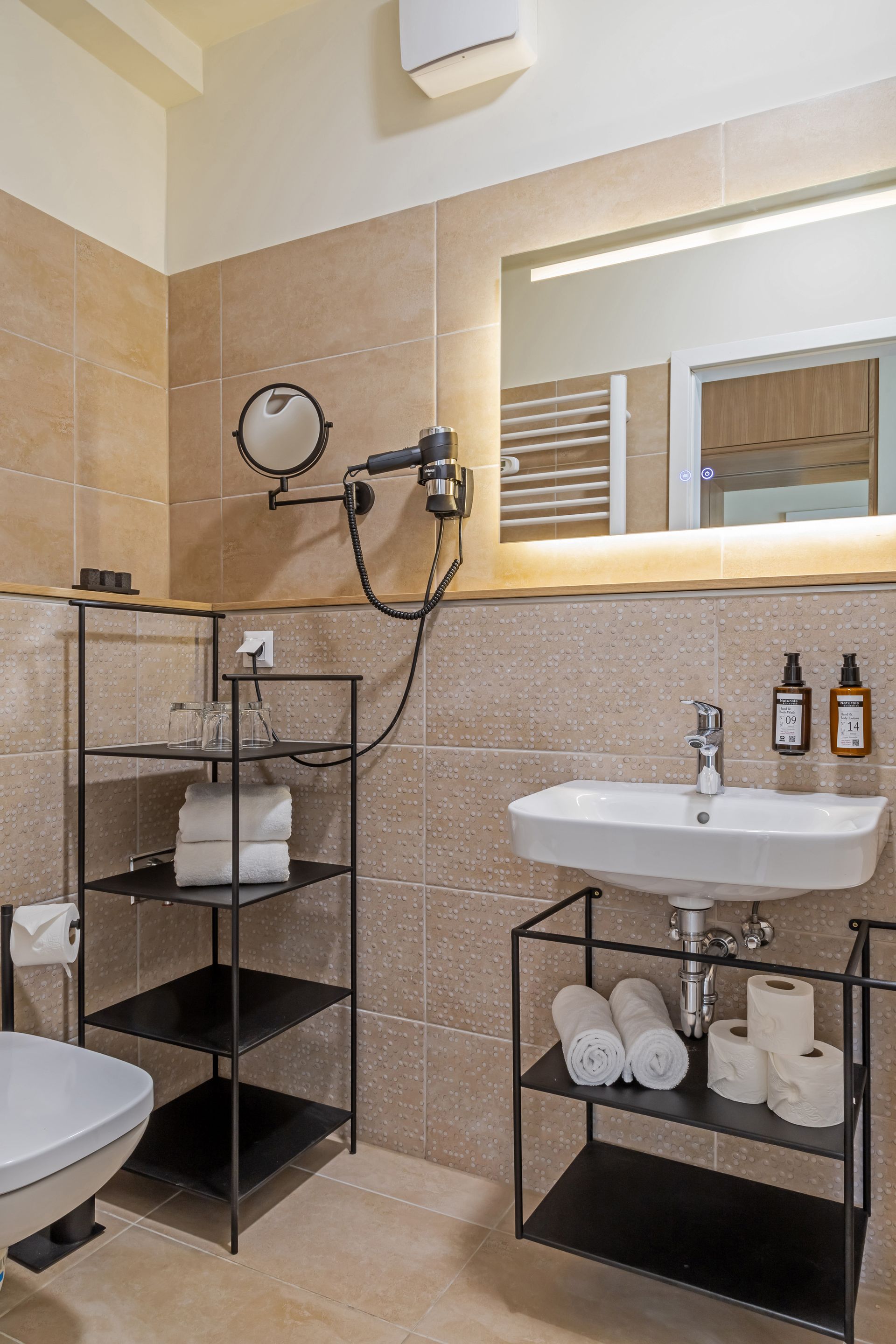 Bathroom with tan tiled walls, black shelving unit with white towels, soap and body cream, and a large mirror. Marone Suites