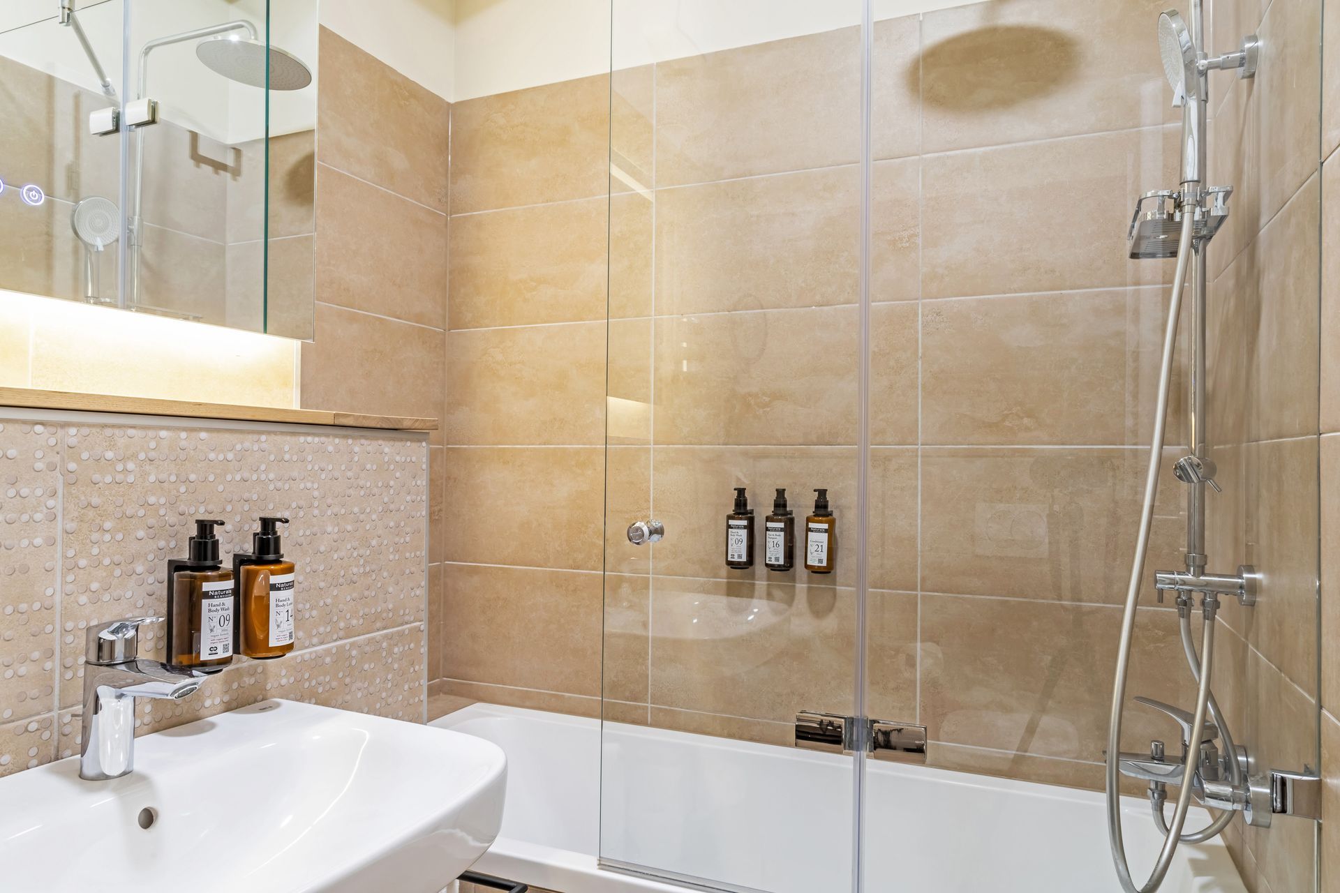 Bathroom with beige tiled walls, white sink, shower and bathtub, glass door, toiletries and white towels in Marone Suites.