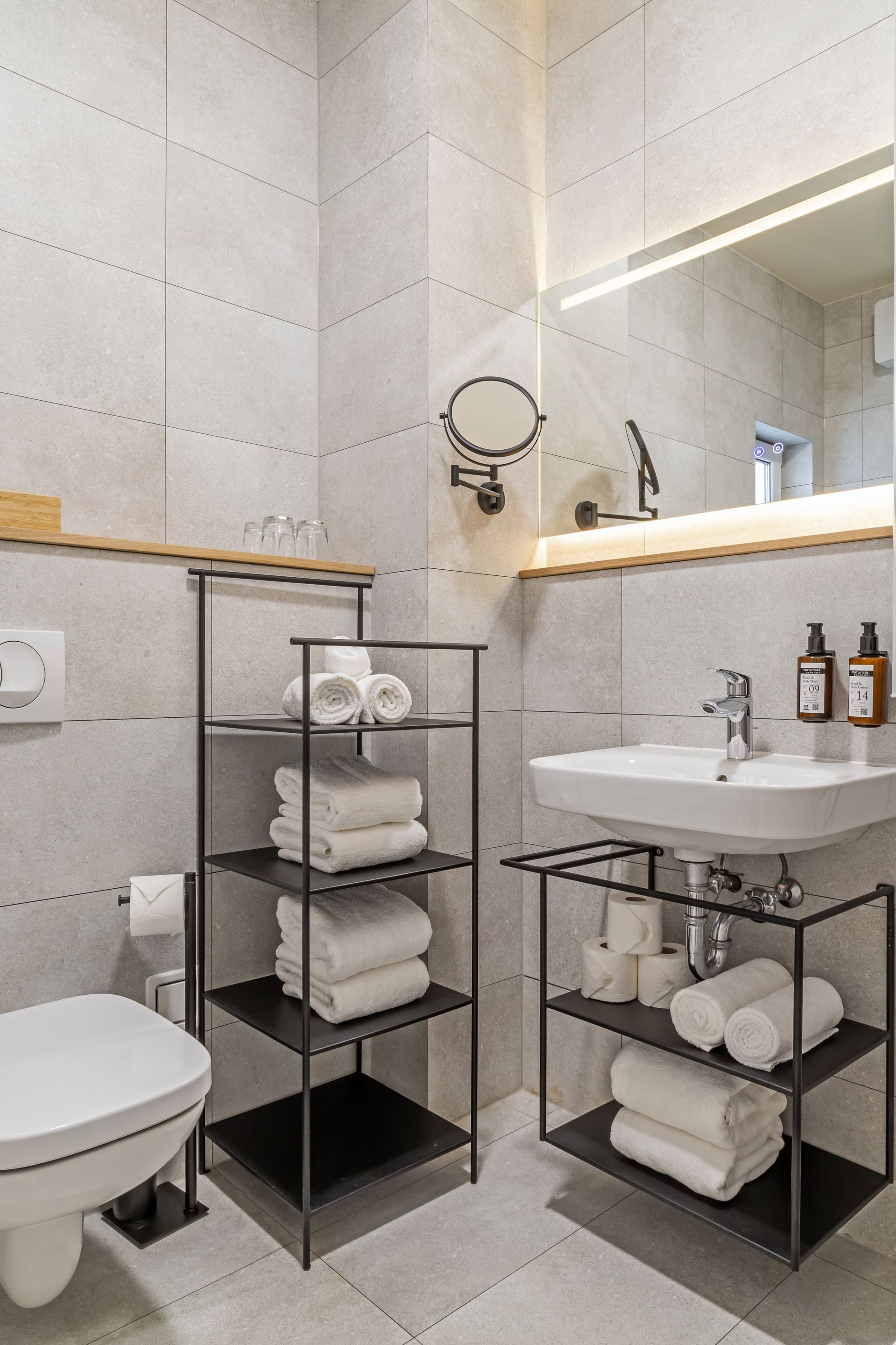 Modern bathroom with white and gray tile, featuring a toilet, sink, toiletries and metal shelving with towels. Marone Suites