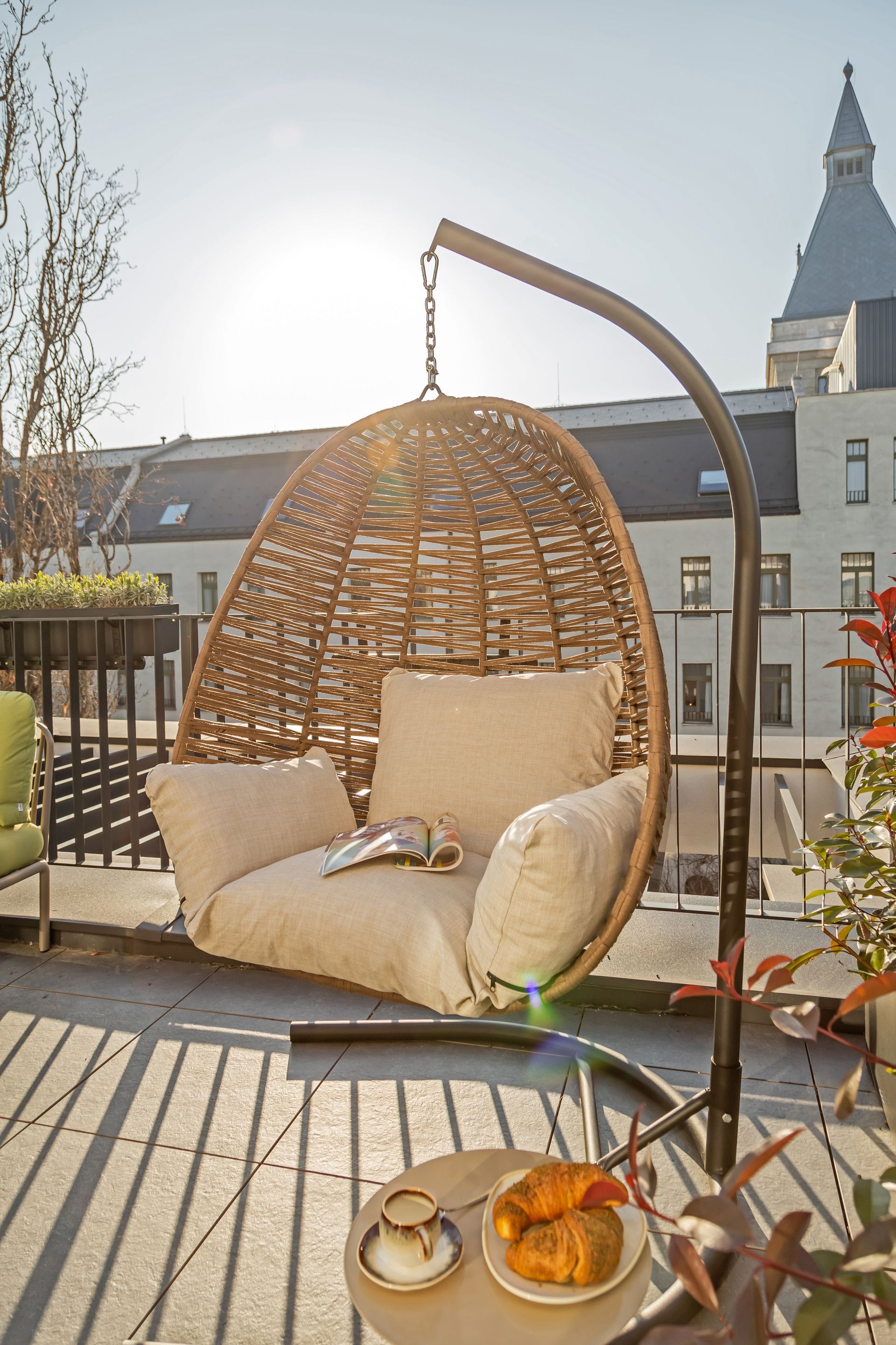 Rooftop balcony with a hanging wicker chair, cushion, side table with coffee and croissants; sunny day in Marone Suites.