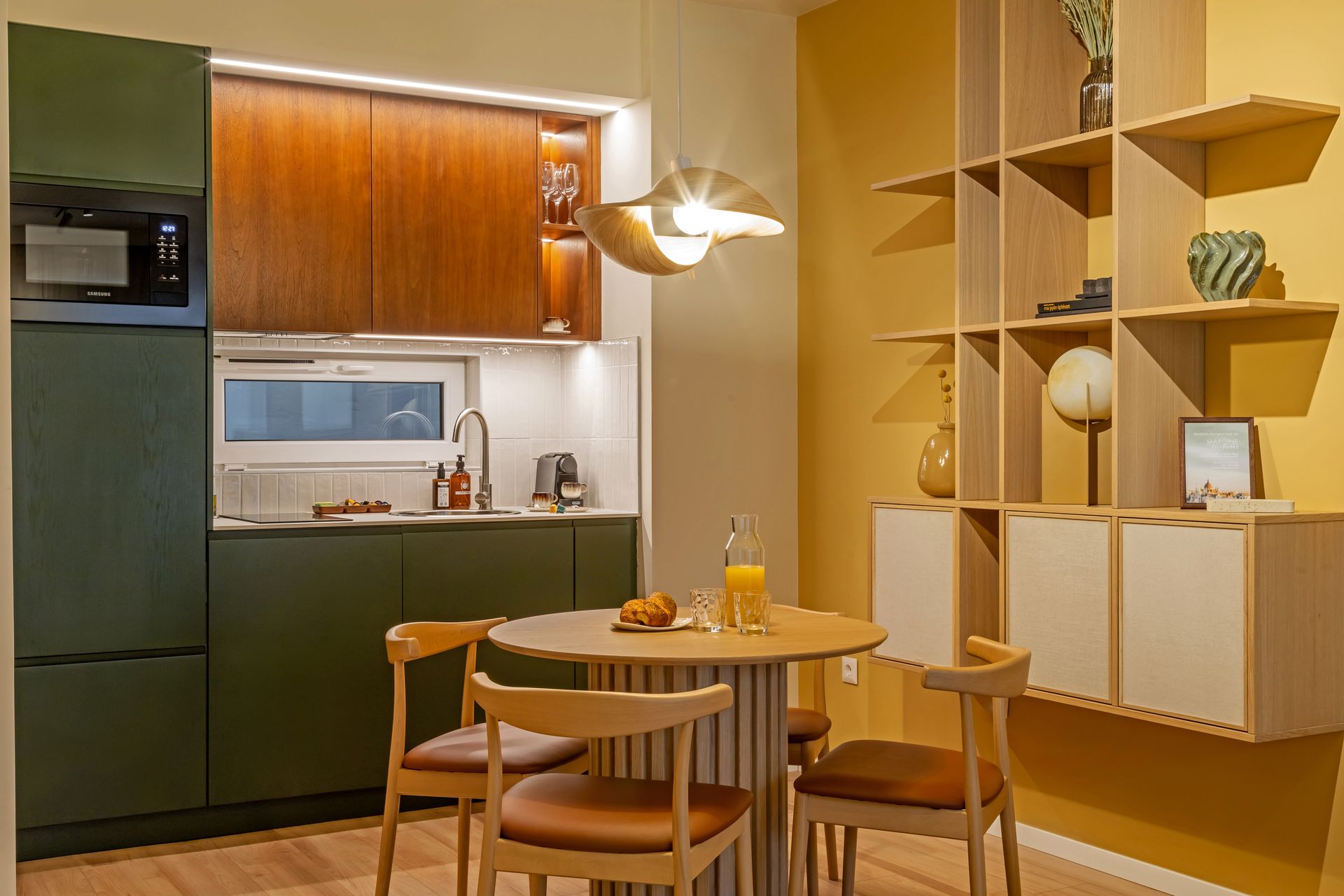 A kitchen with wood and olive green cabinets. A round table with chairs, and a display shelf on the right in Marone Suites.